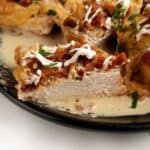 Close-up of a breaded, sliced chicken breast on a plate, topped with ranch sauce, crispy bacon pieces, and fresh herbs.