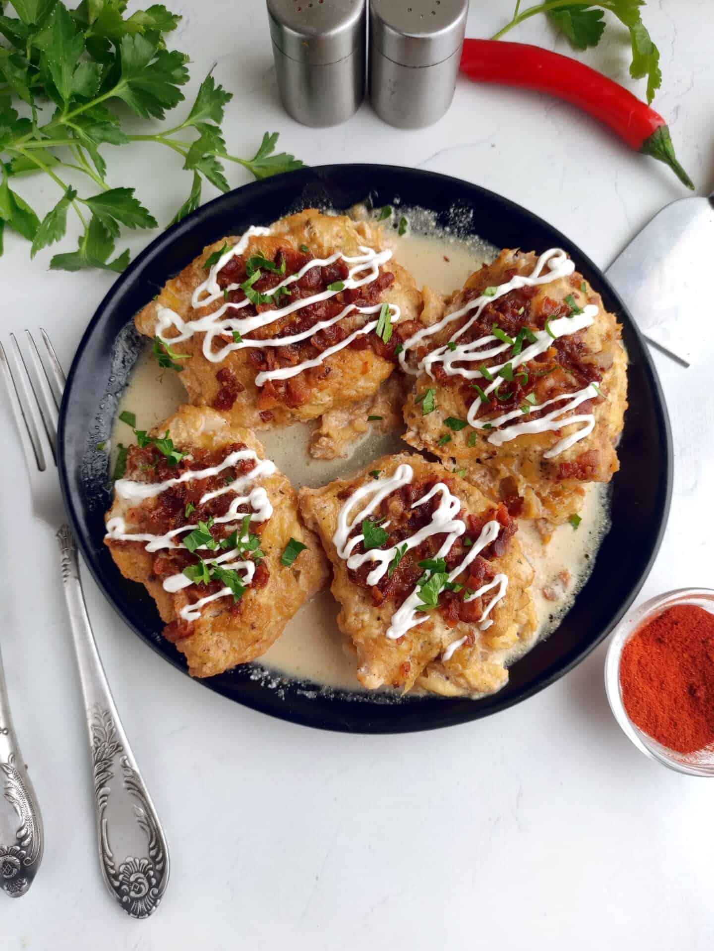 Four breaded Ranch Chicken pieces rest on a black plate, topped with sauce and herbs. Silver utensils, parsley, chili, paprika, and salt shakers surround the plate on a pristine white surface.