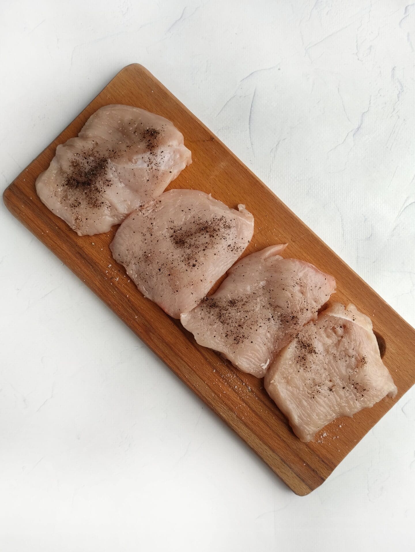Four pieces of raw chicken breast seasoned with black pepper and a hint of ranch sit on a wooden cutting board.