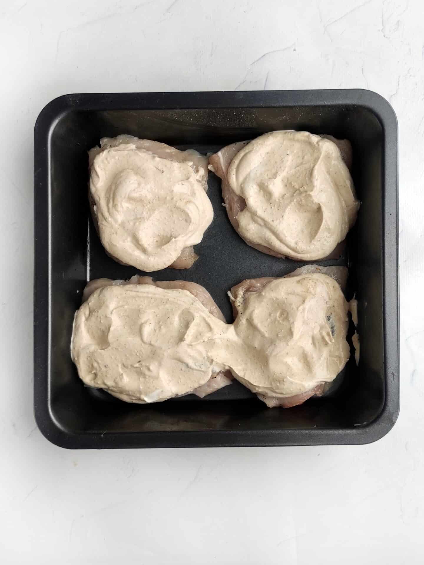 Four pieces of Ranch Chicken topped with a creamy white sauce, nestled in a black baking dish on a pristine white surface.