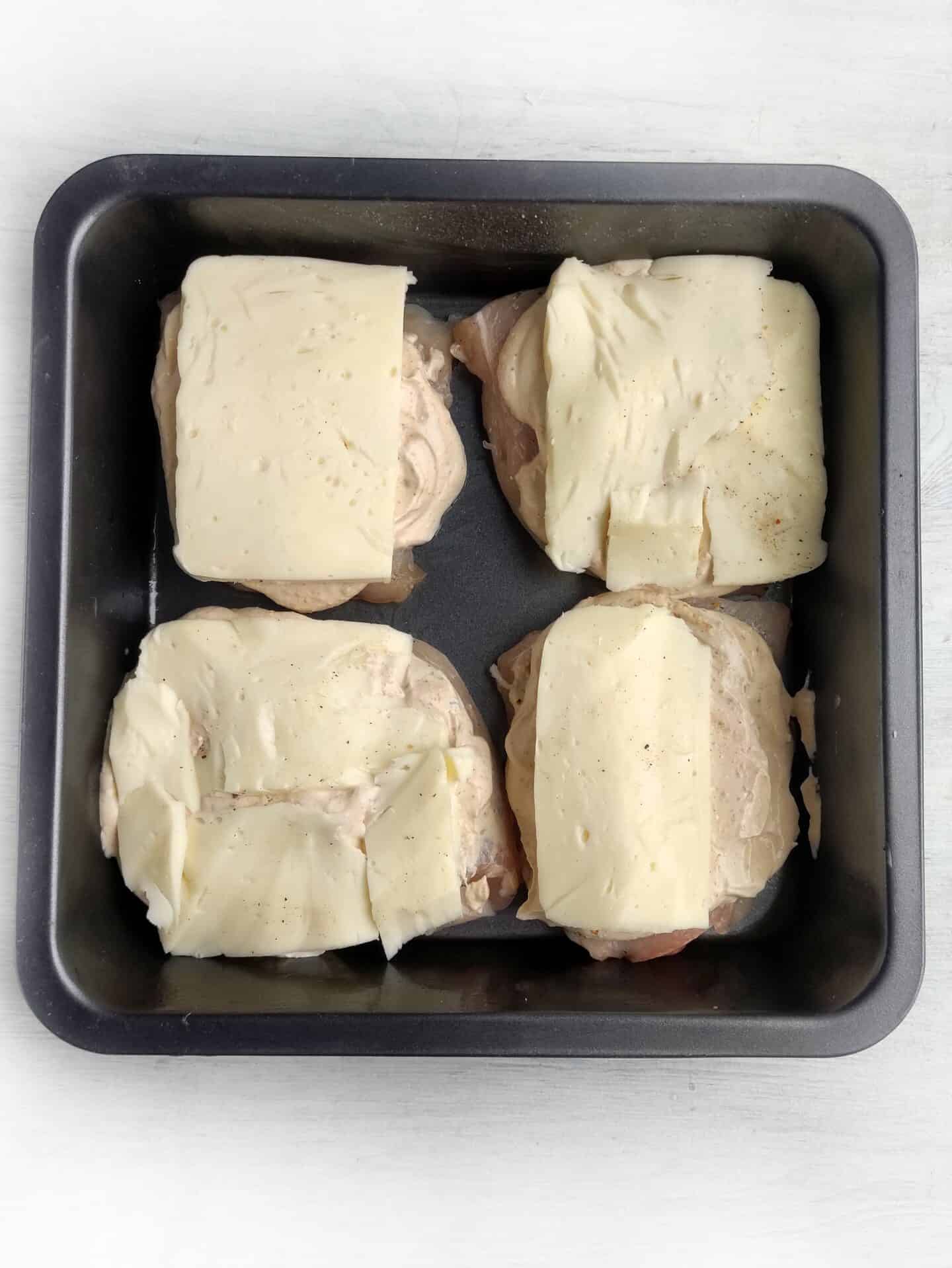 Four pieces of chicken in a baking tray, each topped with a slice of cheese, have been seasoned to perfection.