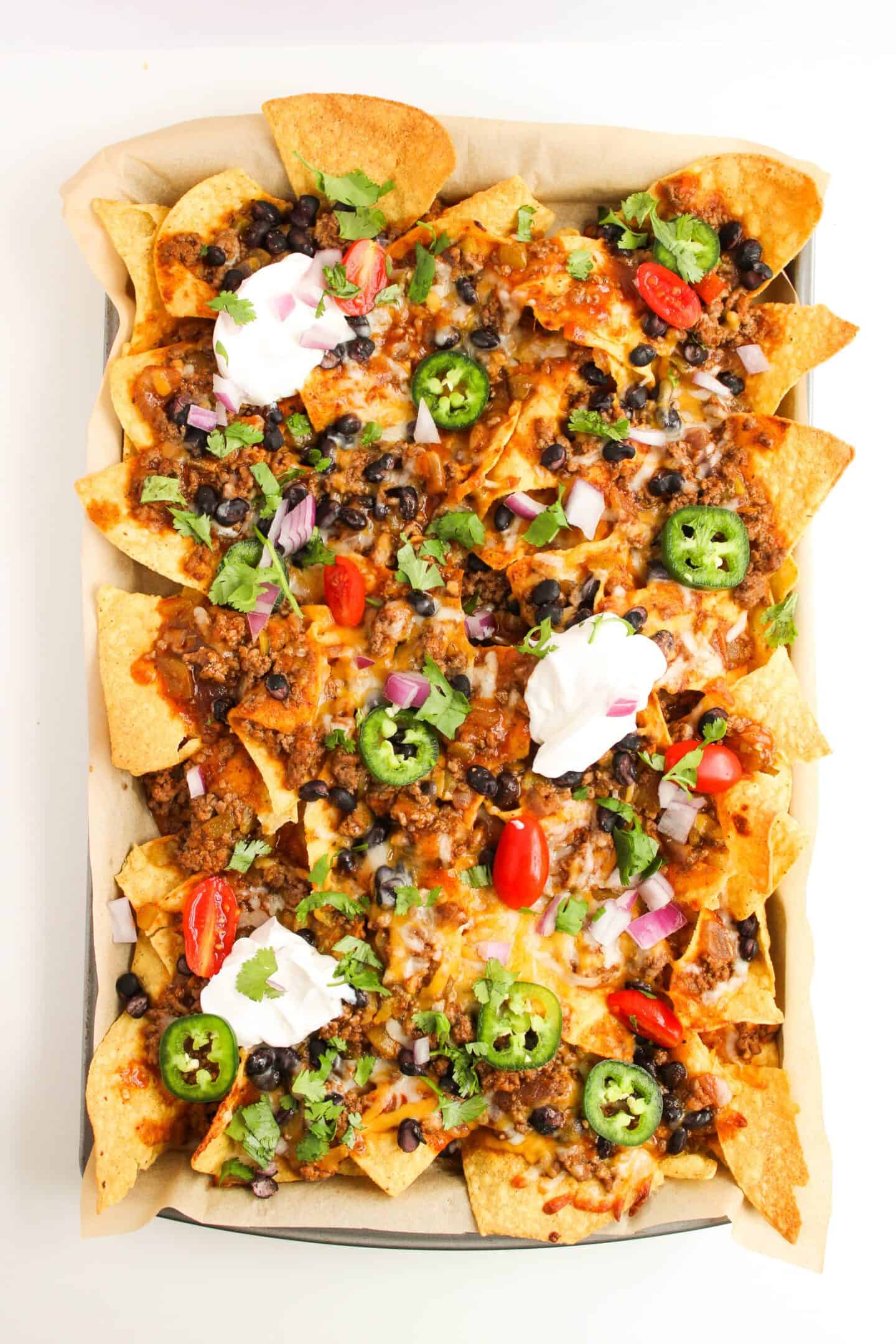 A sheet pan of loaded nachos topped with melted cheese, ground beef, black beans, jalapeños, cherry tomatoes, red onion, cilantro, and dollops of sour cream is reminiscent of delicious Beef Enchilada Nachos.