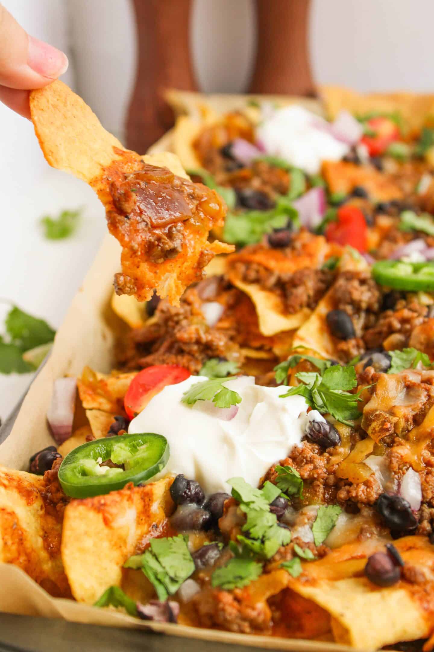 A close-up of Beef Enchilada Nachos with toppings like ground meat, cheese, beans, sliced jalapeños, sour cream, and cilantro. A hand holds a chip topped with melted cheese and meat from the sheet pan.