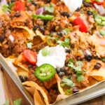 A sheet pan piled with beef enchilada nachos, topped with ground beef, tomatoes, jalapeños, black beans, onions, cilantro, melted cheese, and dollops of sour cream.