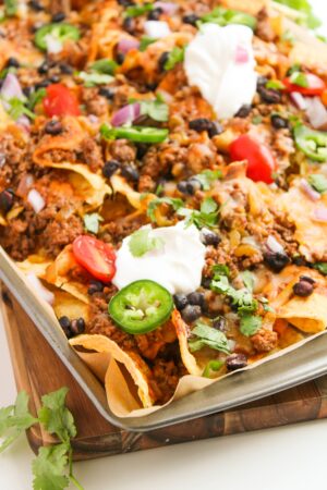 A sheet pan piled with beef enchilada nachos, topped with ground beef, tomatoes, jalapeños, black beans, onions, cilantro, melted cheese, and dollops of sour cream.