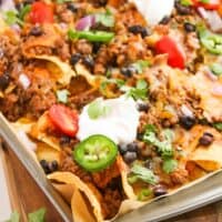 A sheet pan piled with beef enchilada nachos, topped with ground beef, tomatoes, jalapeños, black beans, onions, cilantro, melted cheese, and dollops of sour cream.