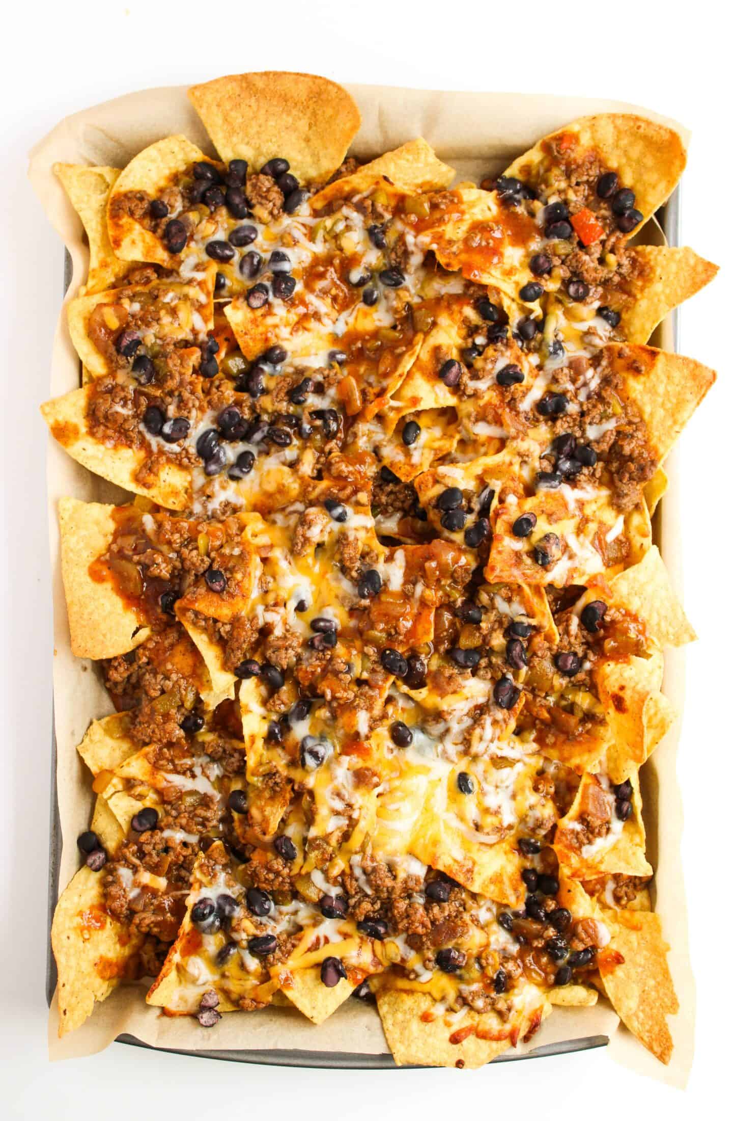 A sheet pan of nachos is generously topped with melted cheese, seasoned ground beef, black beans, and salsa for the ultimate Beef Enchilada Nachos experience.