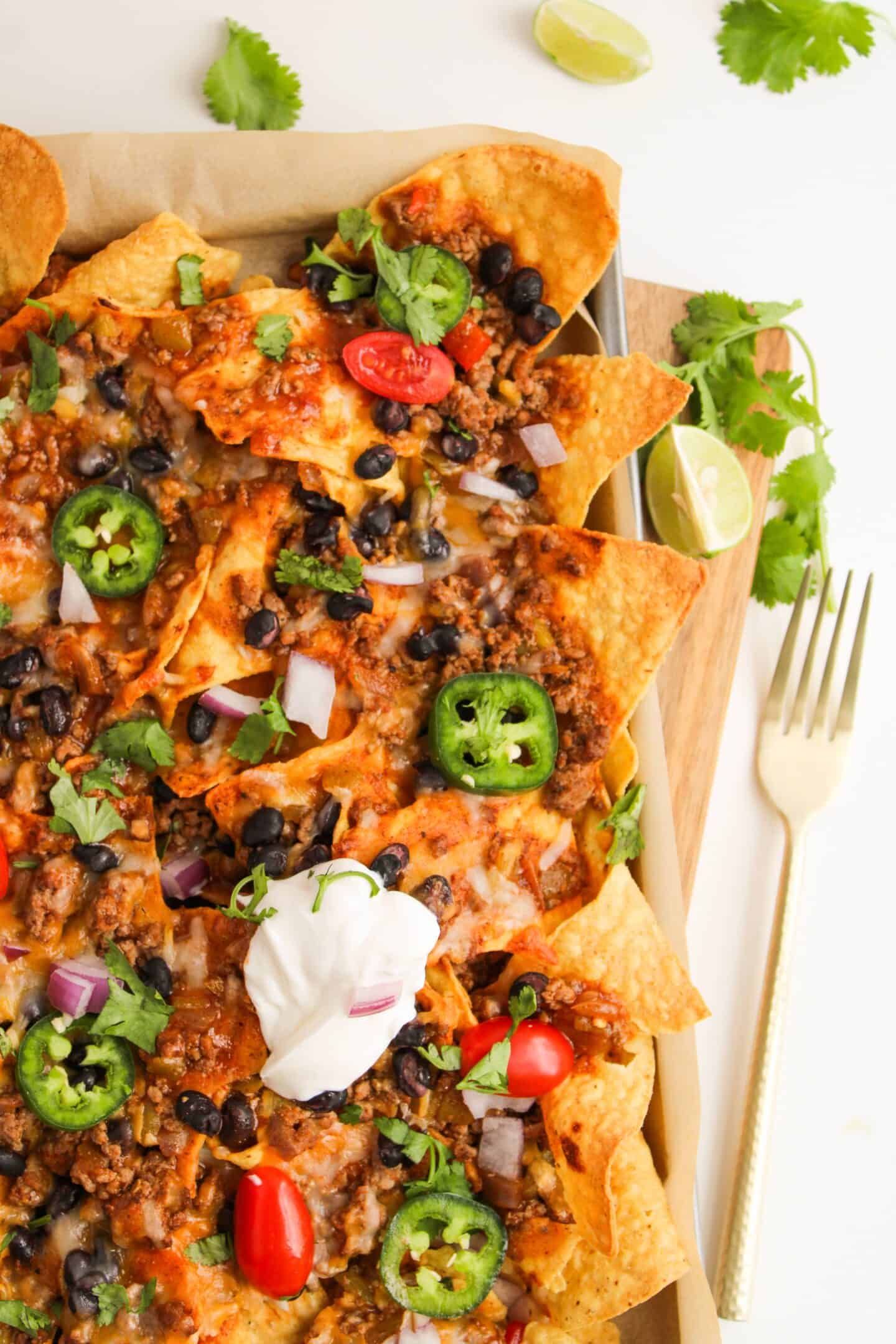 A sheet pan of loaded nachos topped with seasoned ground beef, black beans, melted cheese, jalapeños, ripe tomatoes, red onions, and fresh cilantro. A dollop of sour cream sits on top, perfectly garnished with lime wedges and extra cilantro.