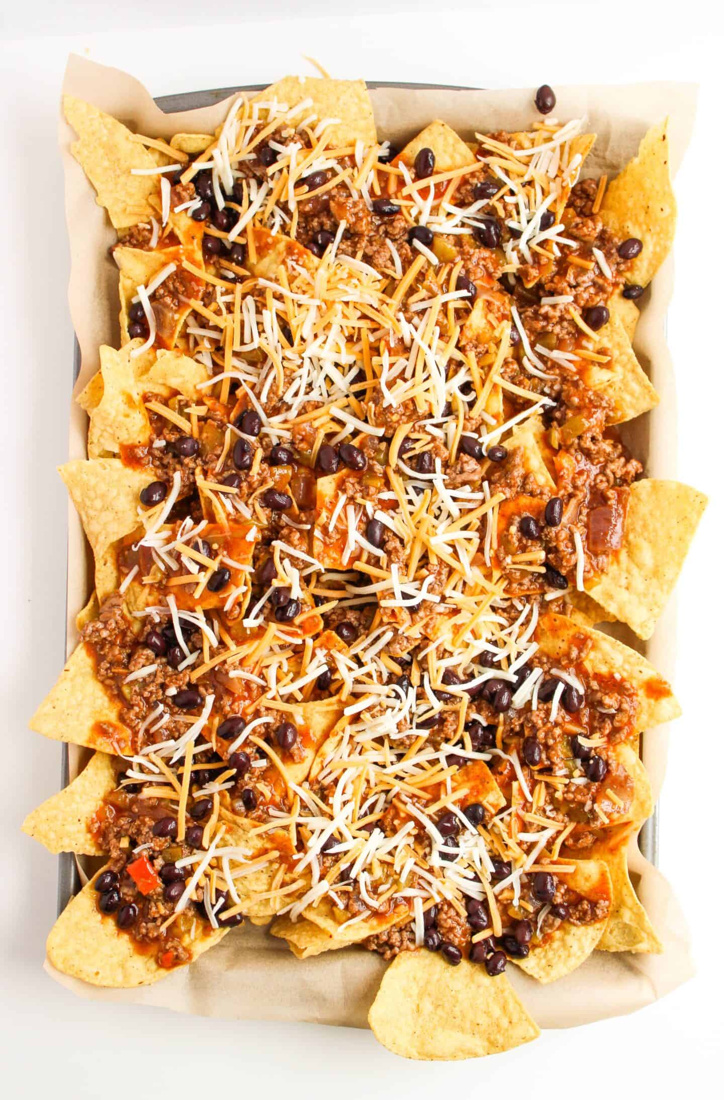 A sheet pan of enchilada nachos topped with ground beef, black beans, diced tomatoes, and shredded cheese.
