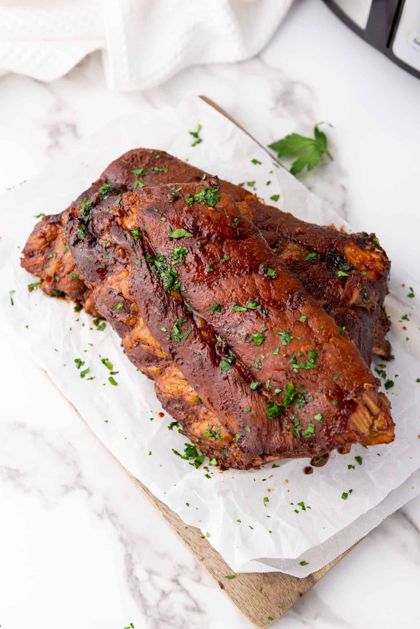 Succulent soy spareribs with a glistening brown glaze, garnished with chopped parsley, placed on parchment paper over a wooden board.