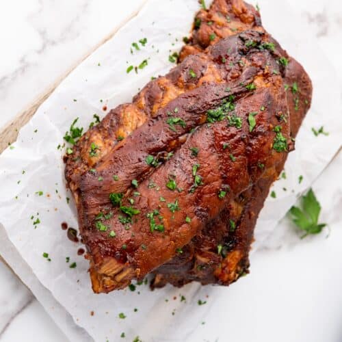 Succulent spareribs, slow-cooked to perfection, are covered in a rich sauce and garnished with chopped parsley on parchment paper, elegantly presented on a marble surface.