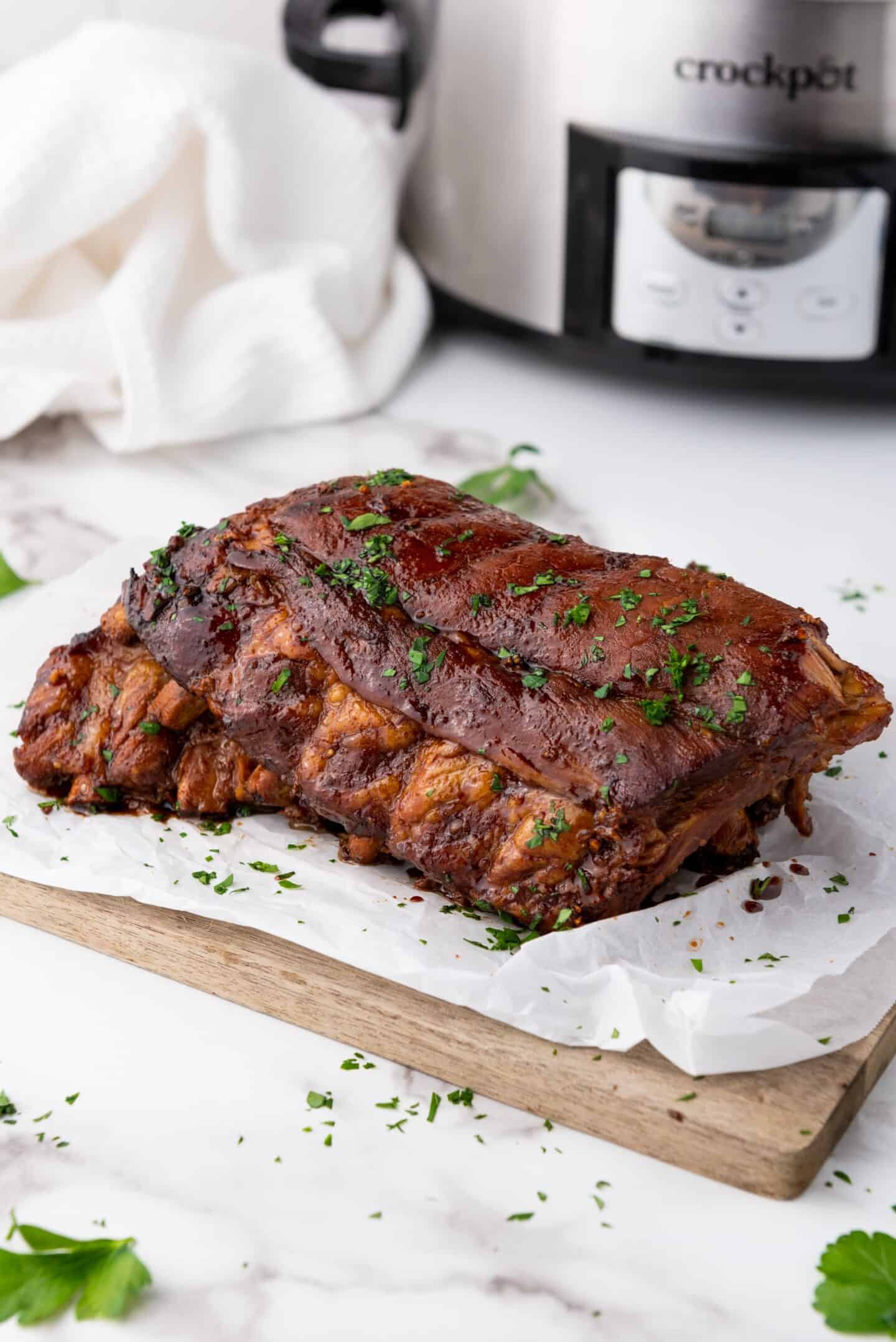 Cooked spareribs garnished with herbs on parchment paper, resting on a wooden board. A slow cooker and white cloth are visible in the background, hinting at the deliciously tender creation process.