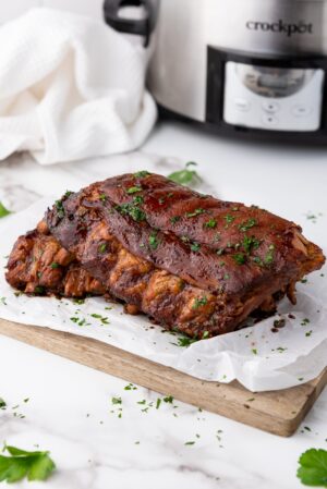 Cooked spareribs garnished with herbs on parchment paper, resting on a wooden board. A slow cooker and white cloth are visible in the background, hinting at the deliciously tender creation process.