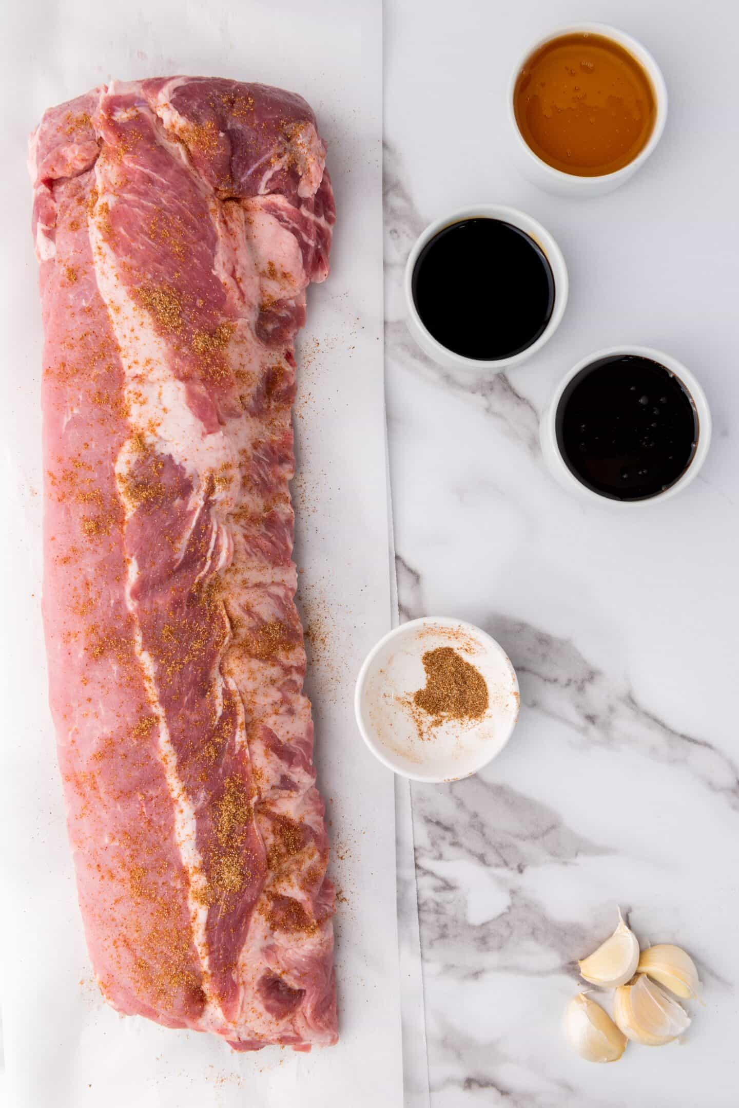 A slab of raw spareribs rests on parchment with a sprinkle of salt, accompanied by small bowls of honey, soy sauce, and seasoning. Garlic cloves scatter across the marble surface, ready to infuse rich flavors in a slow cooker masterpiece.