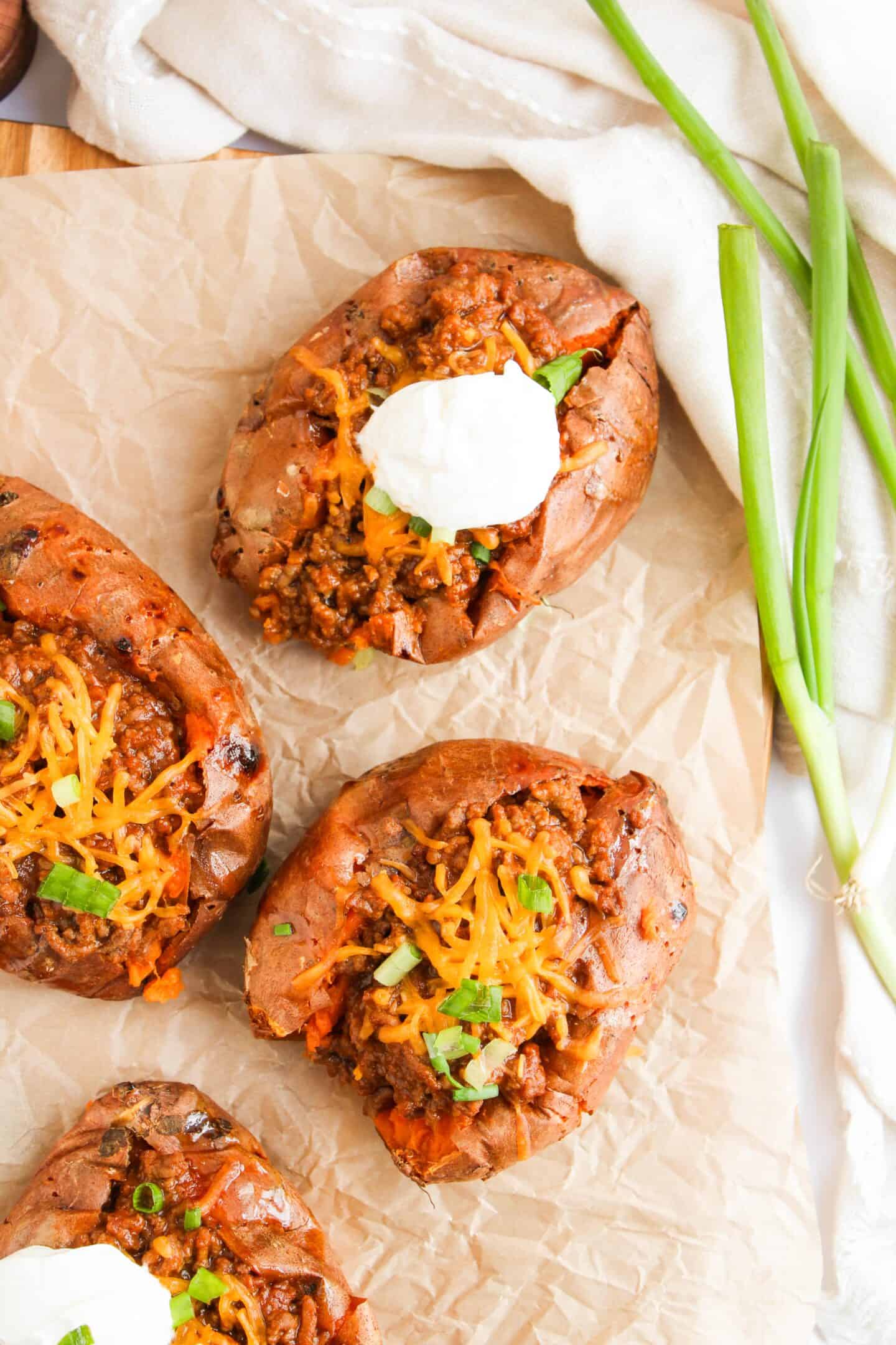 Four sweet potatoes, baked to perfection, are topped with spicy chili, gooey shredded cheese, and a dollop of sour cream. Garnished with vibrant green onions, this dish offers a delightful twist reminiscent of Sloppy Joes.