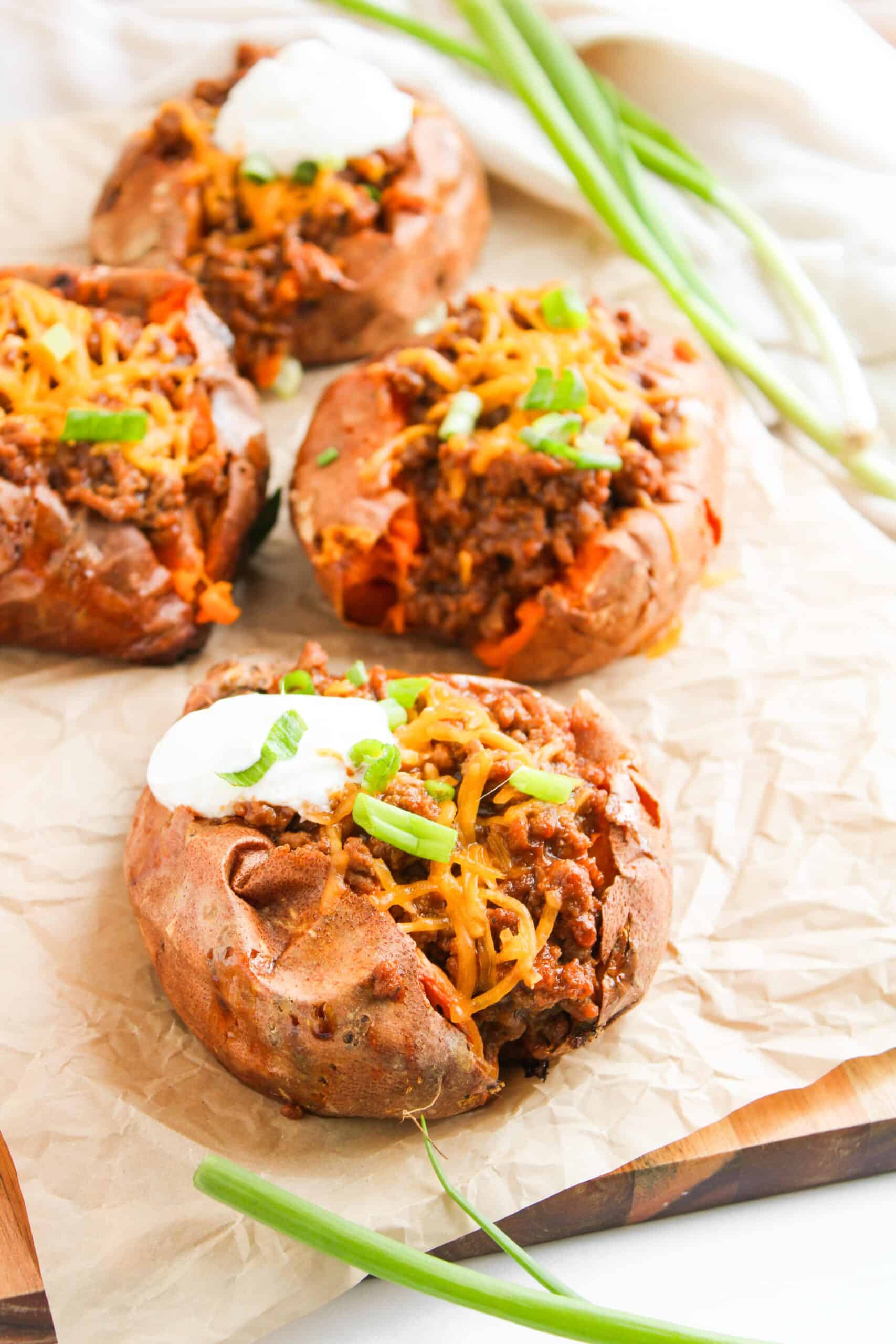 Savory baked sweet potatoes topped with chili, shredded cheese, sour cream, and a sprinkle of green onions, all deliciously assembled on parchment paper.