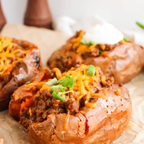 Baked sweet potatoes topped with savory Sloppy Joes, melted cheese, and chopped green onions on parchment paper.
