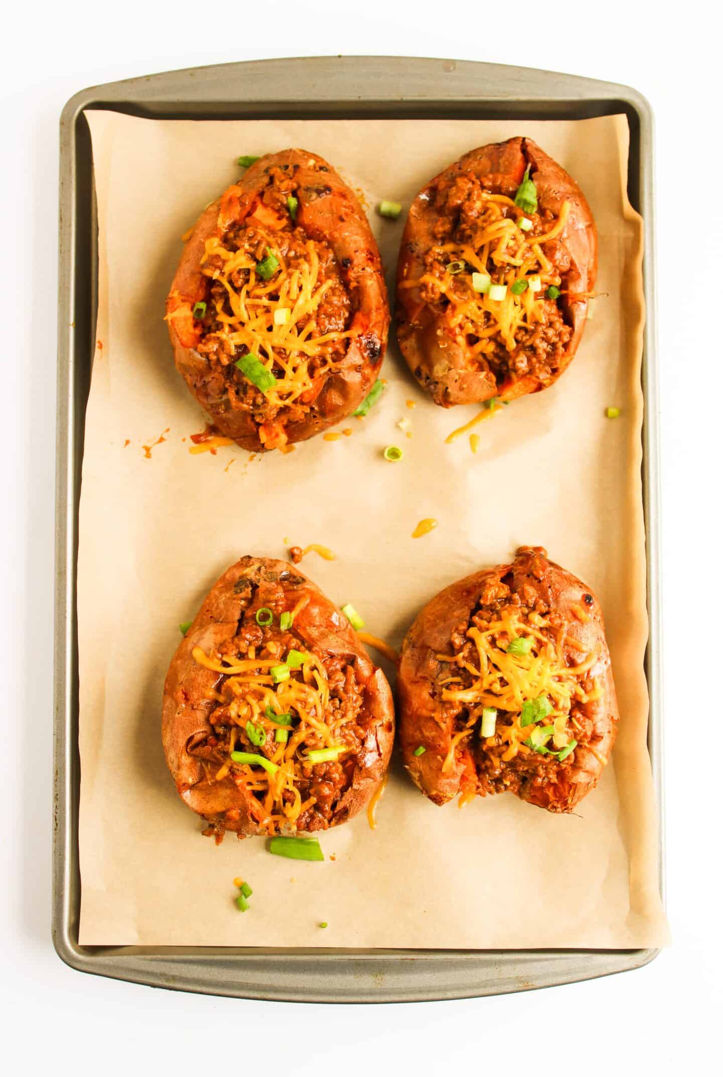 Four baked sweet potatoes topped with a delicious Sloppy Joe mixture, melted cheese, and fresh green onions, all arranged perfectly on a parchment-lined baking tray.