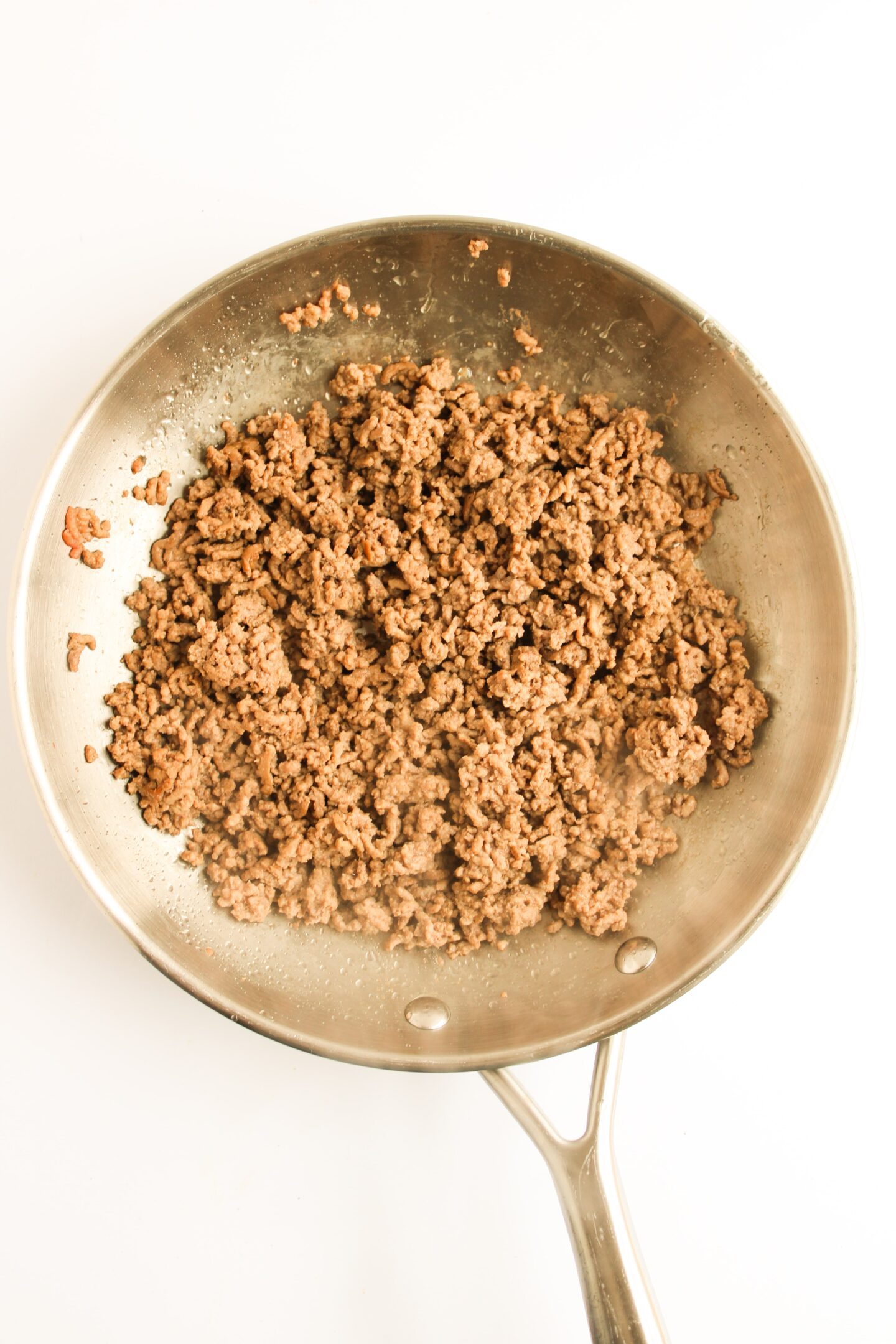 Ground beef sizzles in a stainless steel skillet on a white background, ready to be transformed into delicious Sloppy Joes.