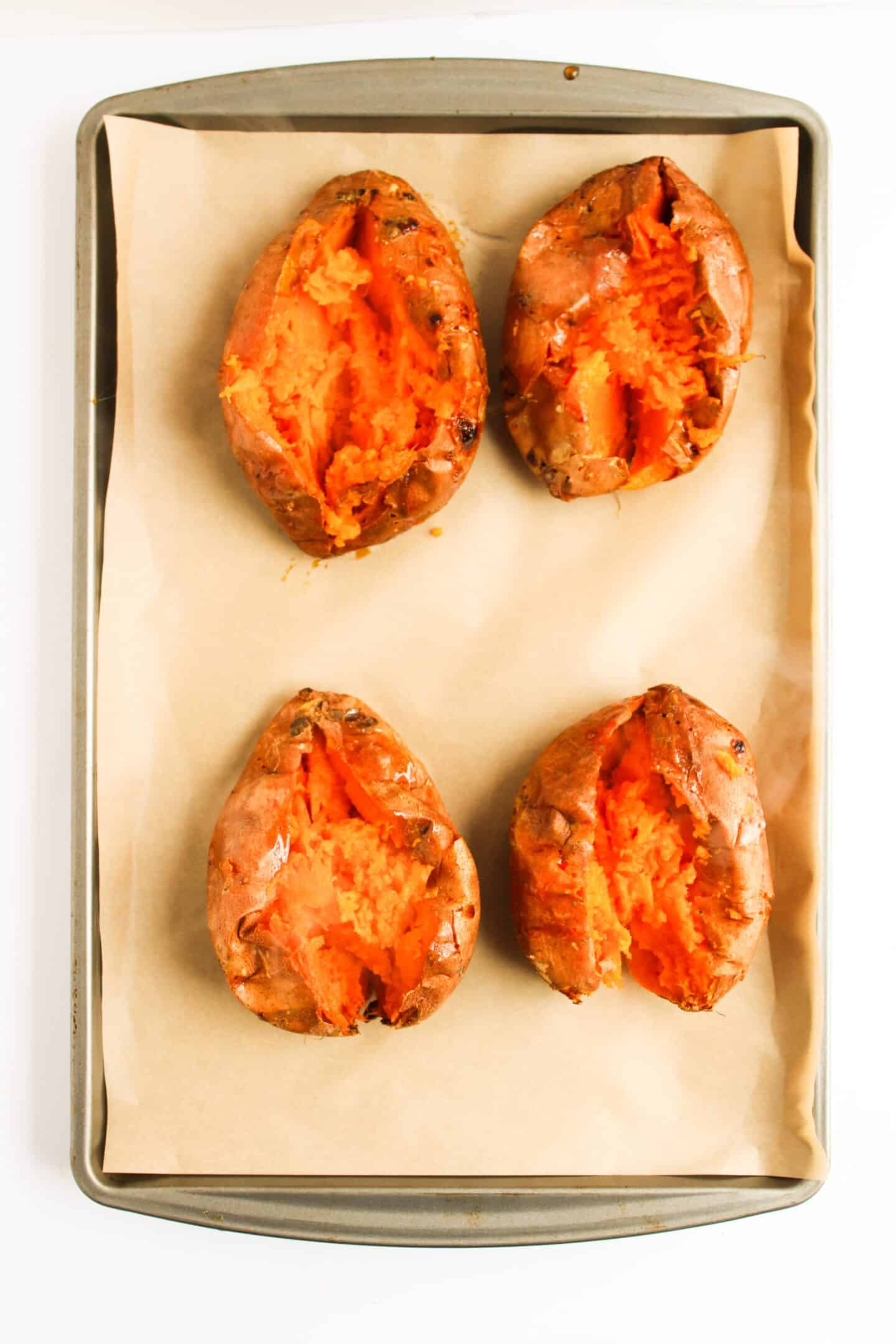 Four baked sweet potatoes on a parchment-lined baking sheet, their tops split open to reveal tender insides. This versatile base is perfect for your sweet potato recipe collection or even as a twist on classic Sloppy Joes.