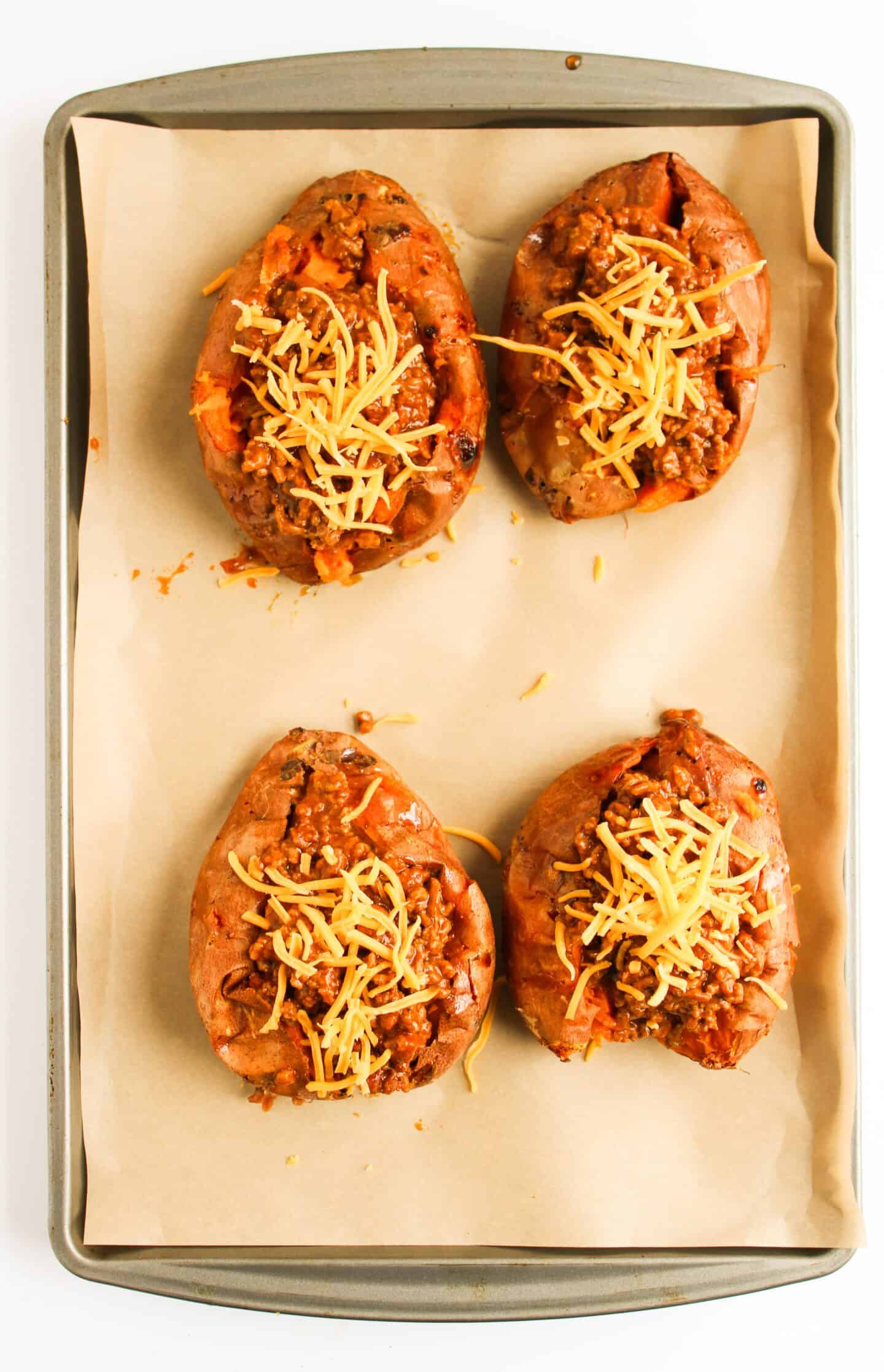 Four baked sweet potatoes infused with the essence of Sloppy Joes, topped generously with ground meat and melted shredded cheese on a parchment-lined baking sheet.
