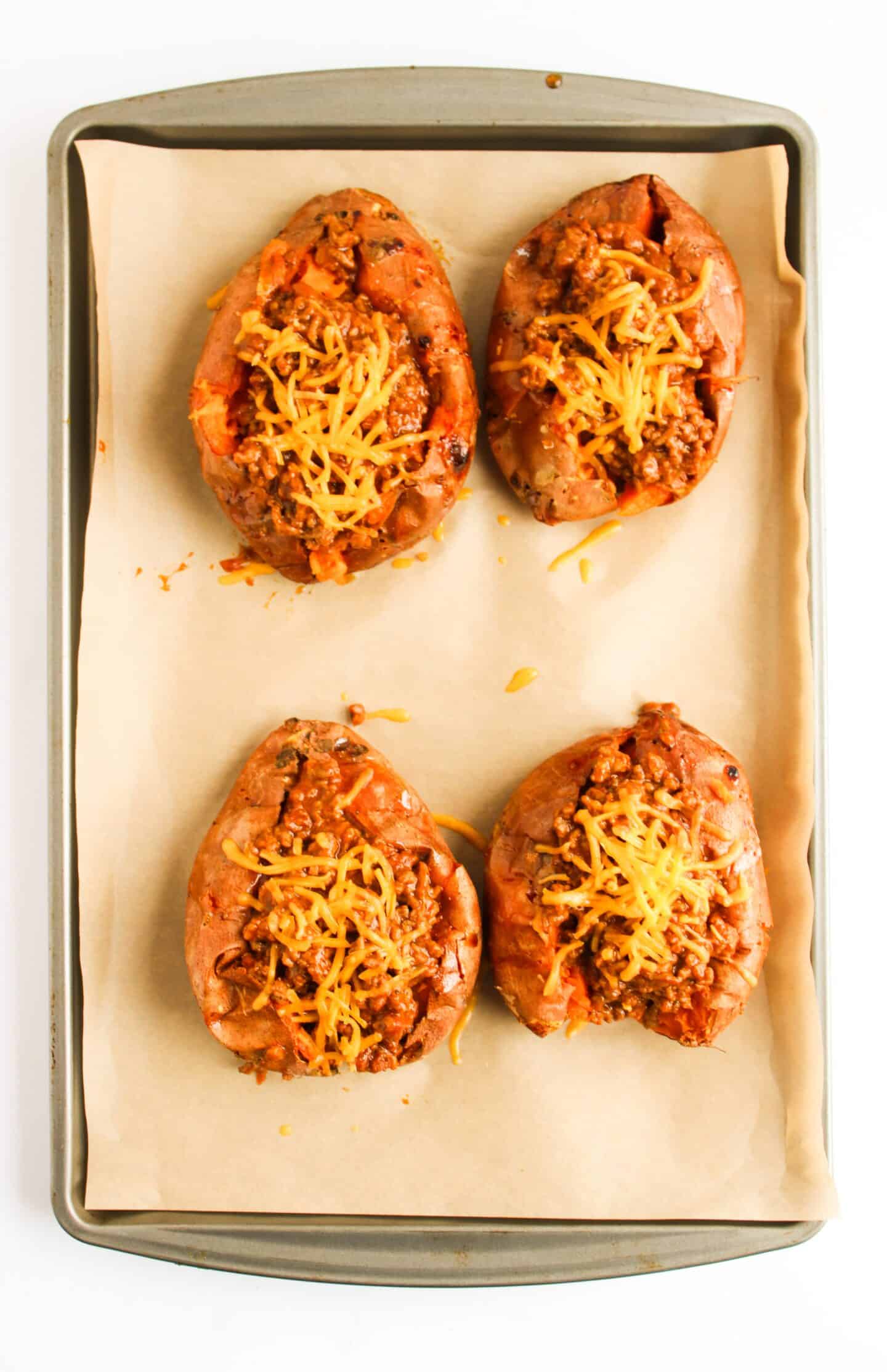 Four baked sweet potatoes loaded with ground meat and melted cheese, reminiscent of Sloppy Joes, all perfectly arranged on a parchment-lined baking sheet.