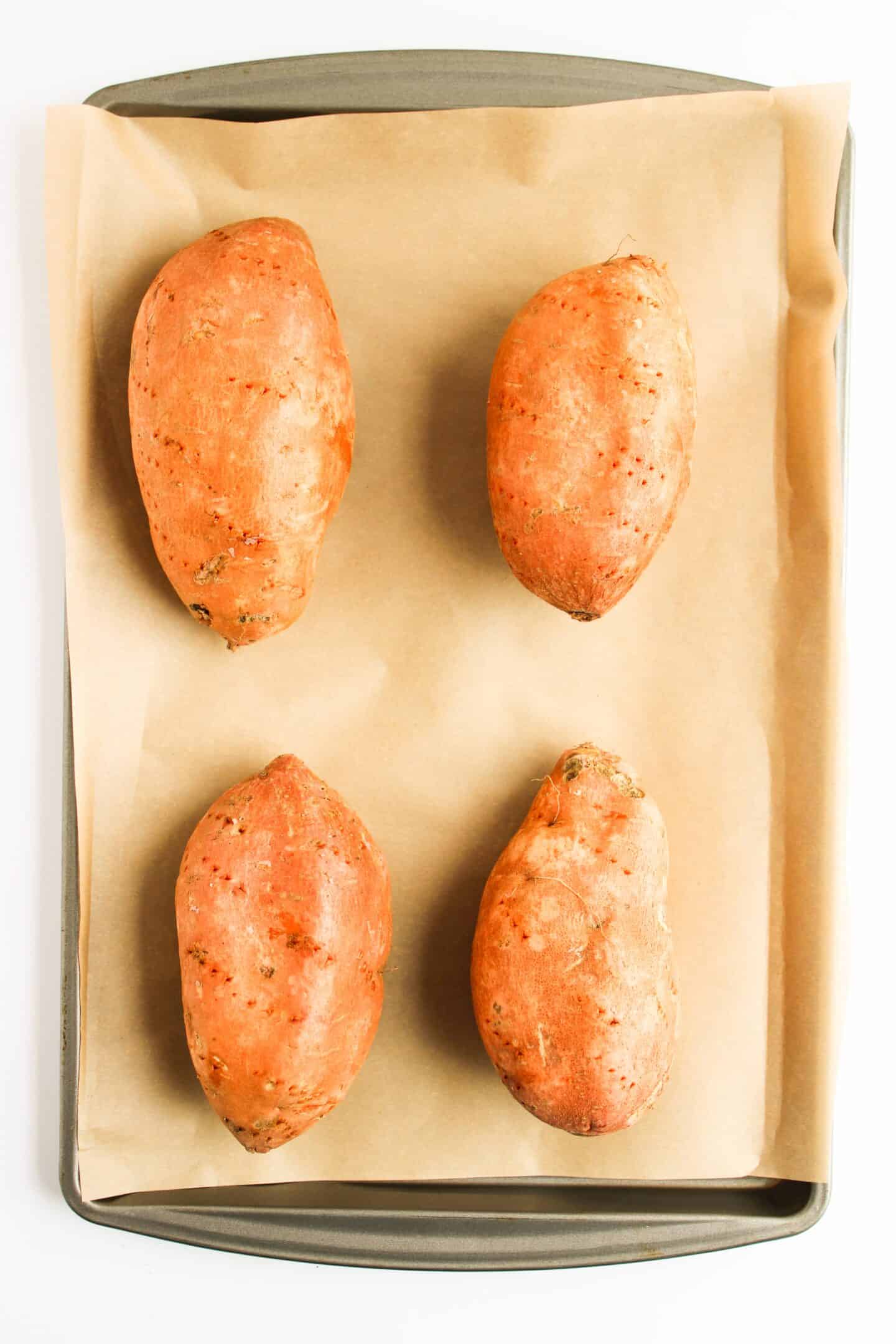 Four whole sweet potatoes sit on parchment paper on a baking sheet, ready to become a delicious twist on classic Sloppy Joes.