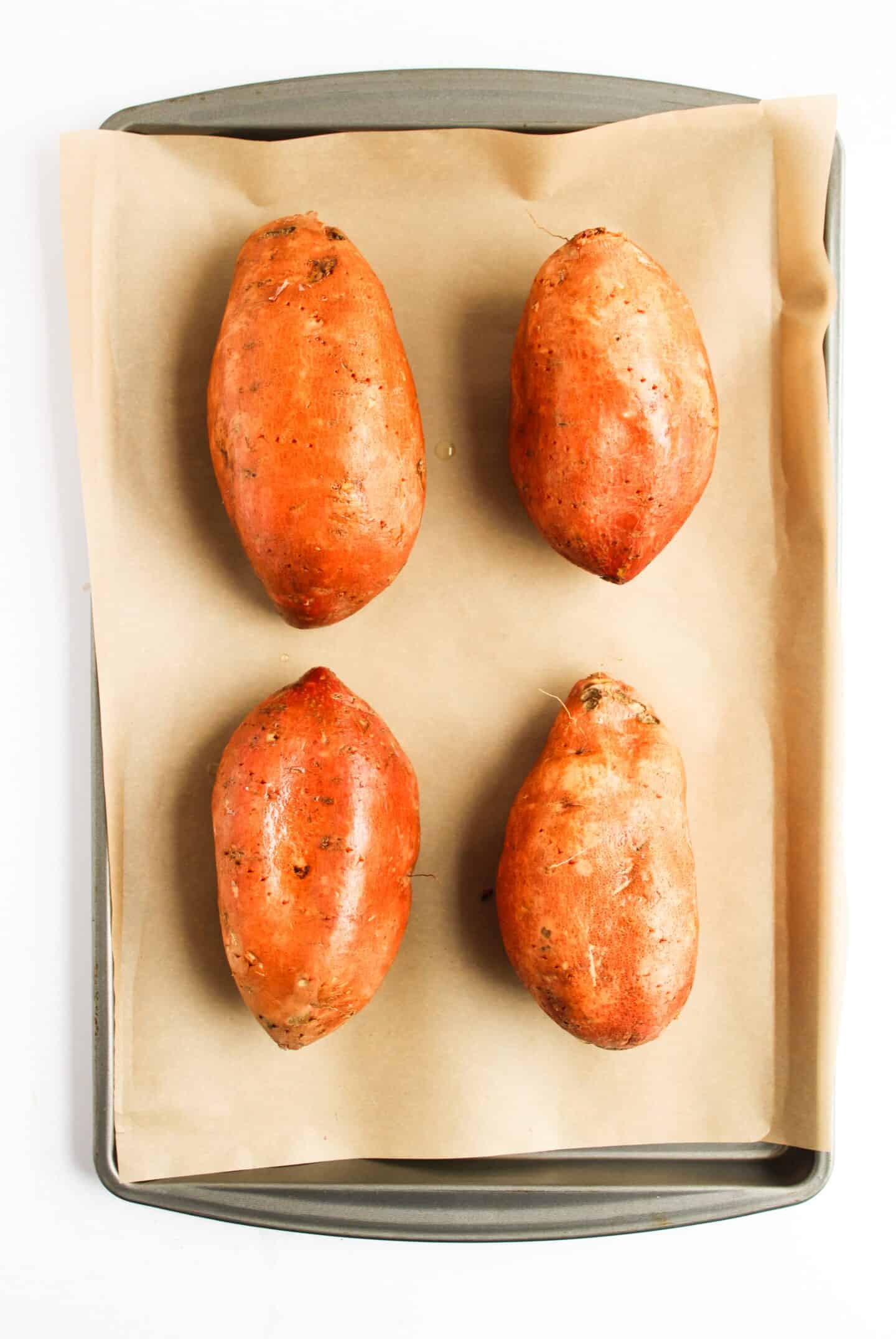 Four whole sweet potatoes rest on a parchment-lined baking tray, ready to be transformed.