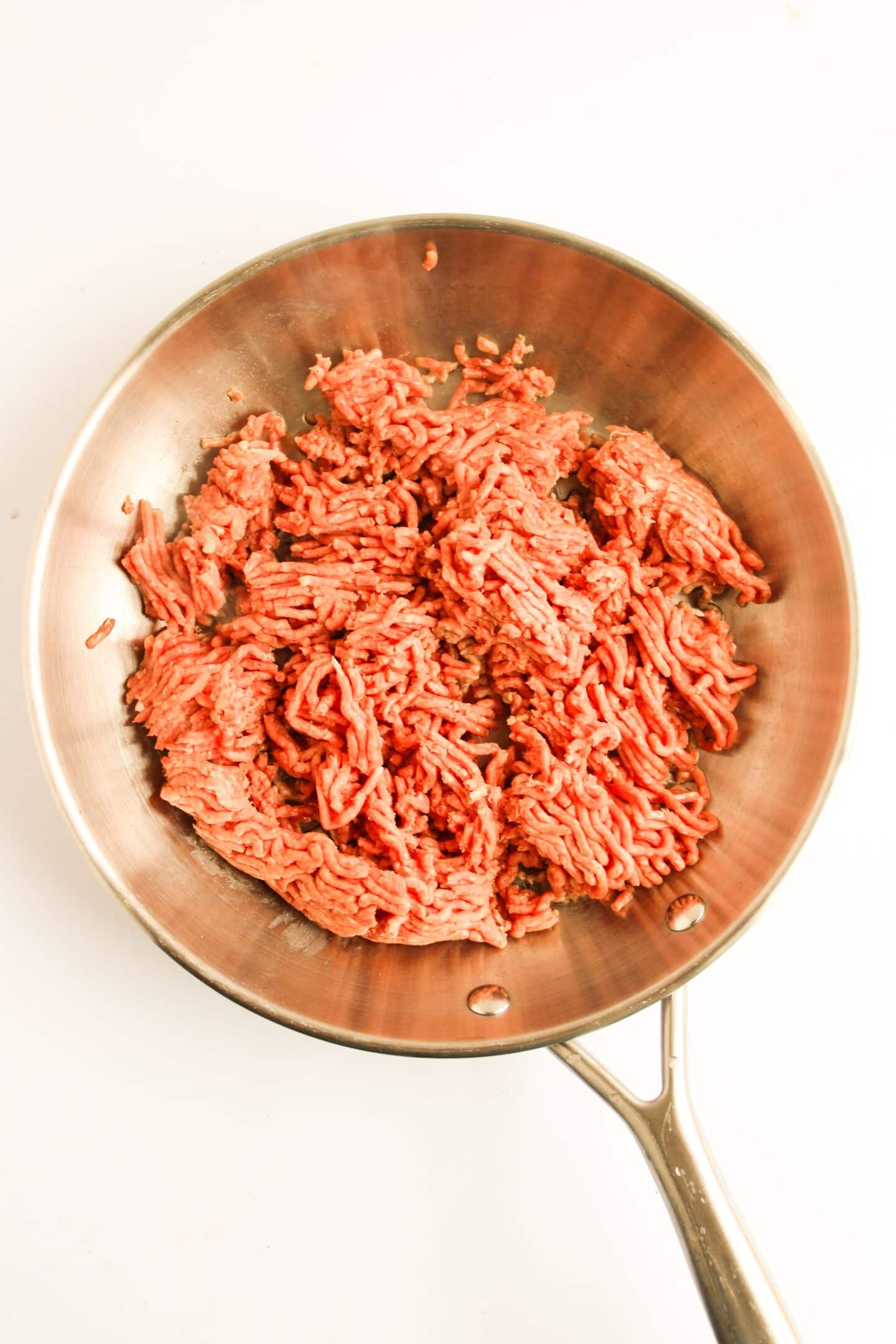 Raw ground beef in a stainless steel frying pan on a white background, ready to be transformed into delightful Sloppy Joes.