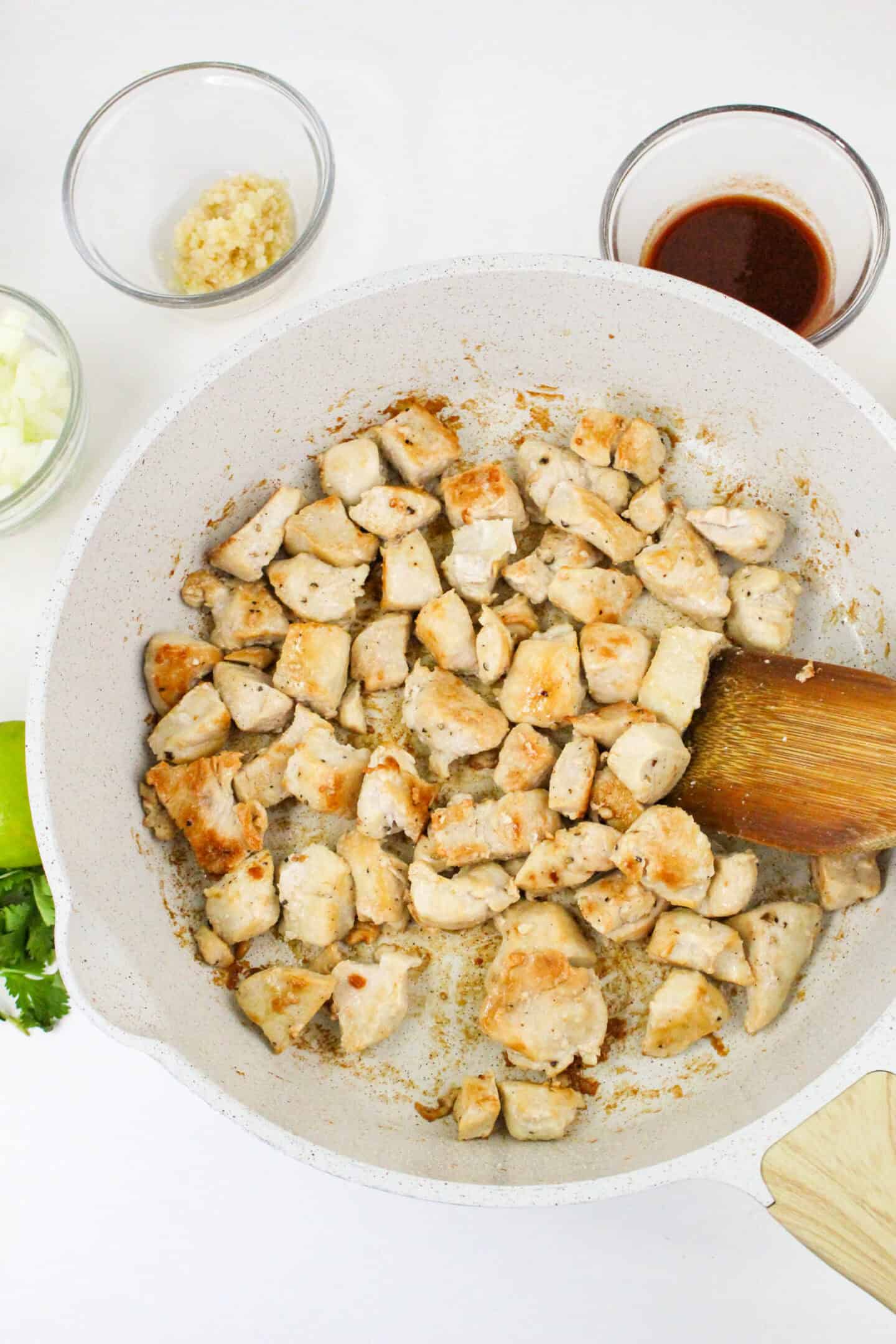 Diced chicken pieces sizzle in a white skillet, stirred with a brown wooden spatula. Nearby, bowls of chopped onions, minced garlic, and sauce await their turn in this soon-to-be chipotle chicken pasta masterpiece on the pristine white surface.