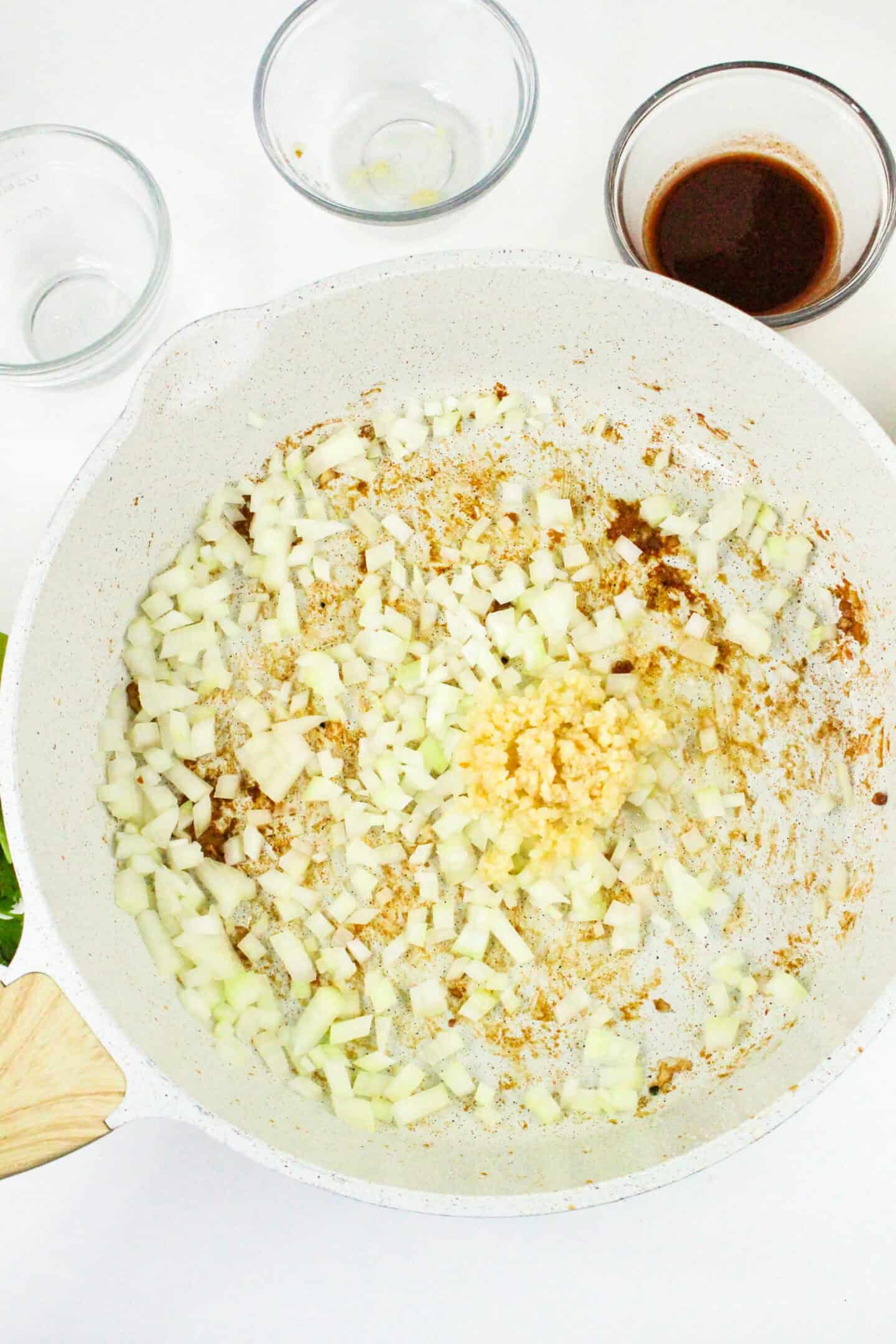 Chopped onions and minced garlic sizzle in a white frying pan, the fragrant base for a savory Chipotle Chicken Pasta. Behind them, two empty glass bowls await their turn alongside a bowl brimming with rich soy sauce.