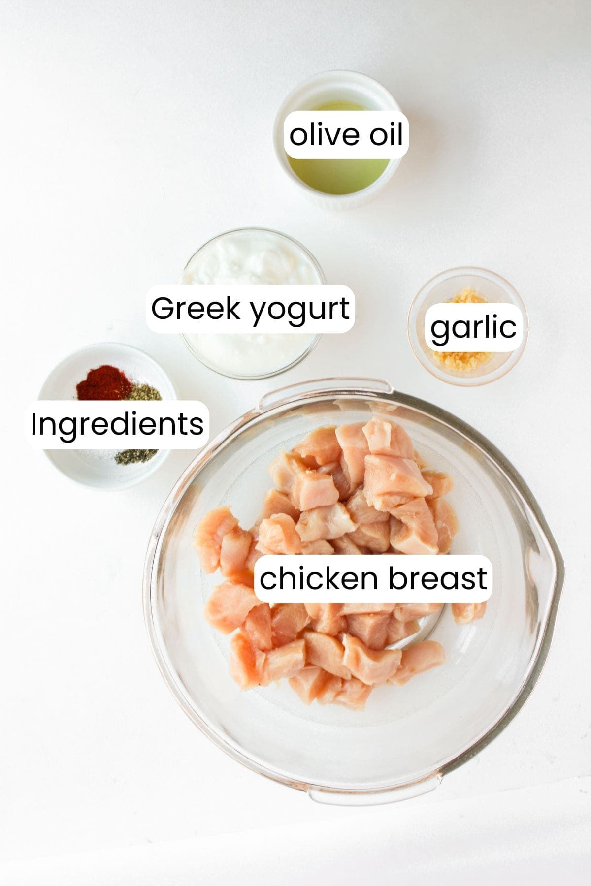 Bowls of diced chicken breast, Greek yogurt dressing, olive oil, minced garlic, and other ingredients on a white surface.