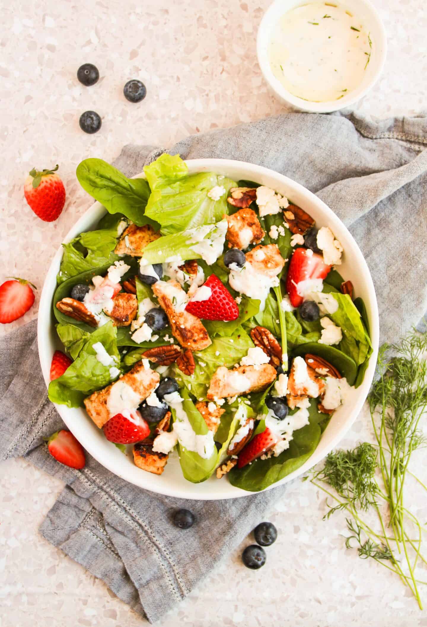 A bowl of vibrant chicken salad featuring spinach, strawberries, blueberries, pecans, and feta. Topped with juicy grilled chicken and a side of dressing for added flavor.