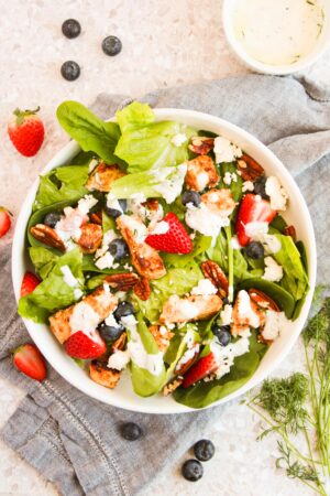 A salad with spinach, strawberries, blueberries, pecans, croutons, and feta cheese is drizzled with a white dressing made creamy with Greek yogurt. It's served in a white bowl on a gray cloth and speckled surface.
