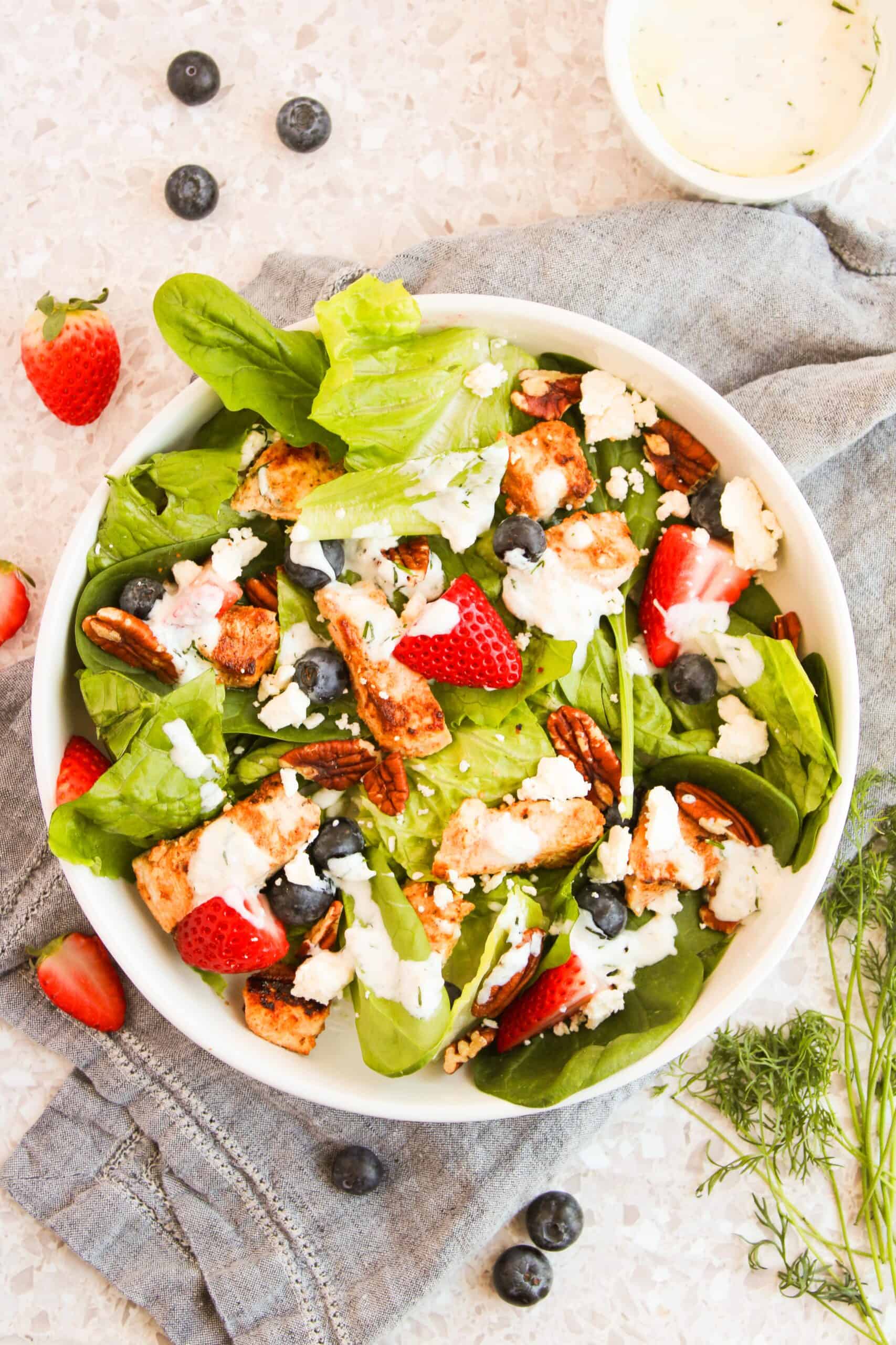 A salad with spinach, strawberries, blueberries, pecans, croutons, and feta cheese is drizzled with a white dressing made creamy with Greek yogurt. It's served in a white bowl on a gray cloth and speckled surface.