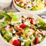 Two bowls of chicken salad featuring grilled chicken, strawberries, blueberries, pecans, and crumbled cheese. A small dish of Greek yogurt dressing is in the background.