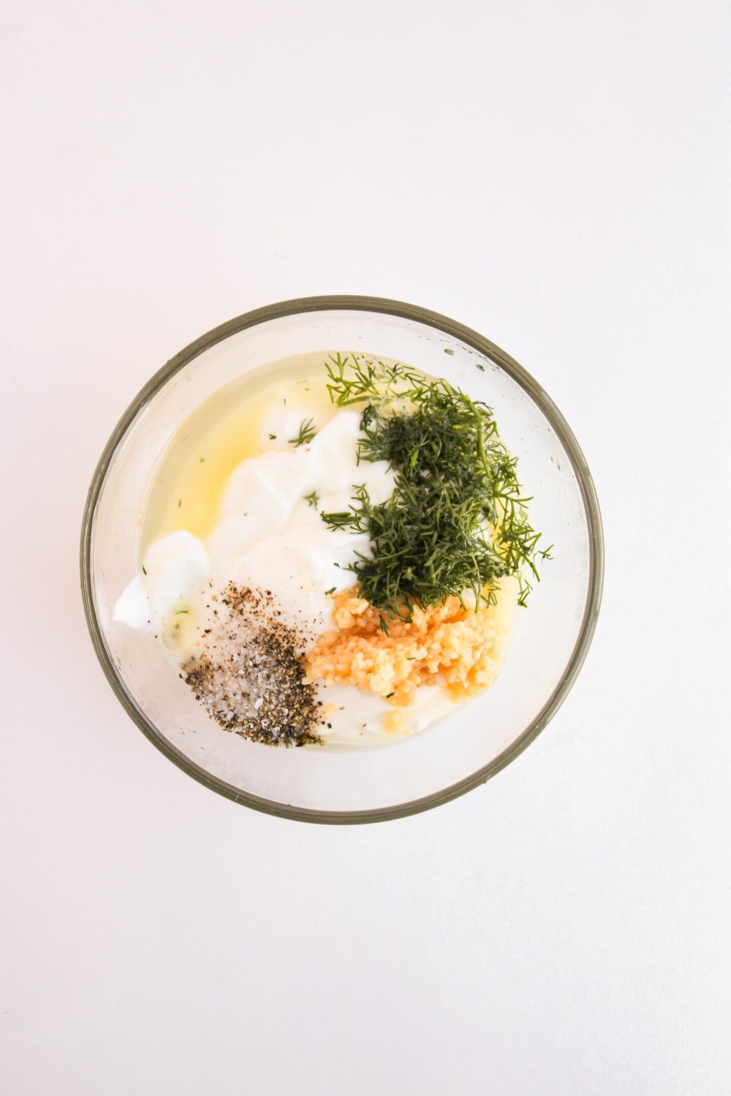 A glass bowl filled with a Greek yogurt dressing—perfect for chicken salad—combines minced garlic, dill, black pepper, and a drizzle of olive oil, all set against a plain white background.