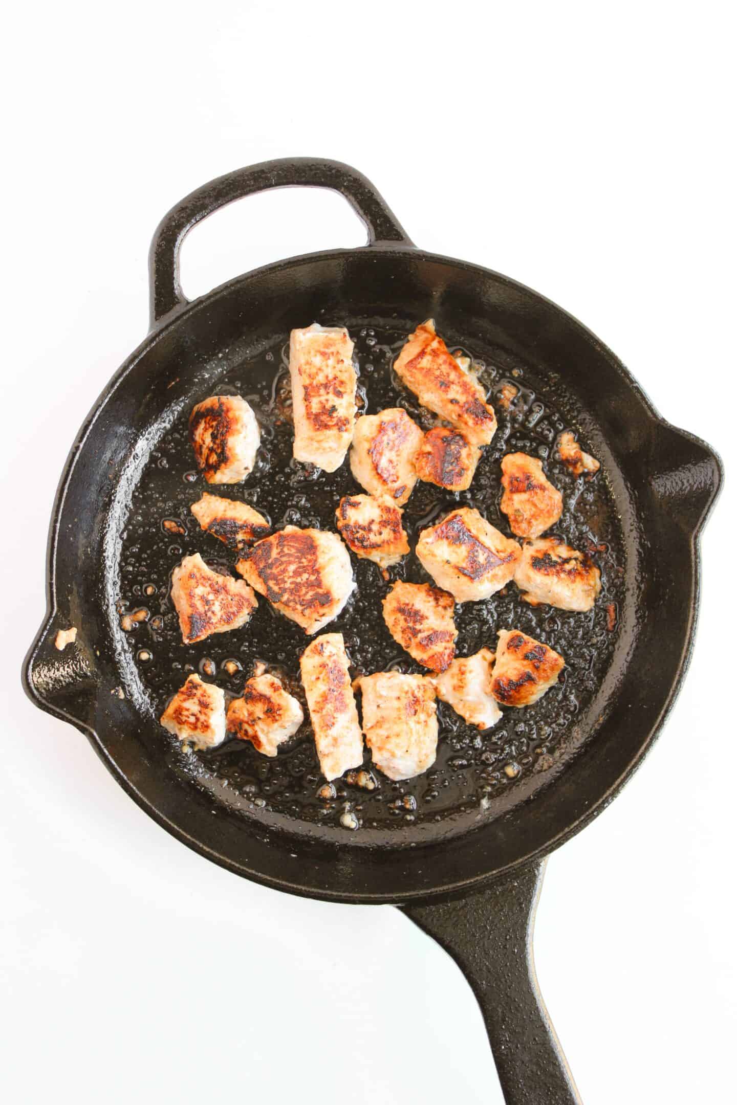 Chunks of cooked chicken, perfect for a chicken salad with Greek yogurt dressing, sit in a black cast-iron skillet against a stark white background.
