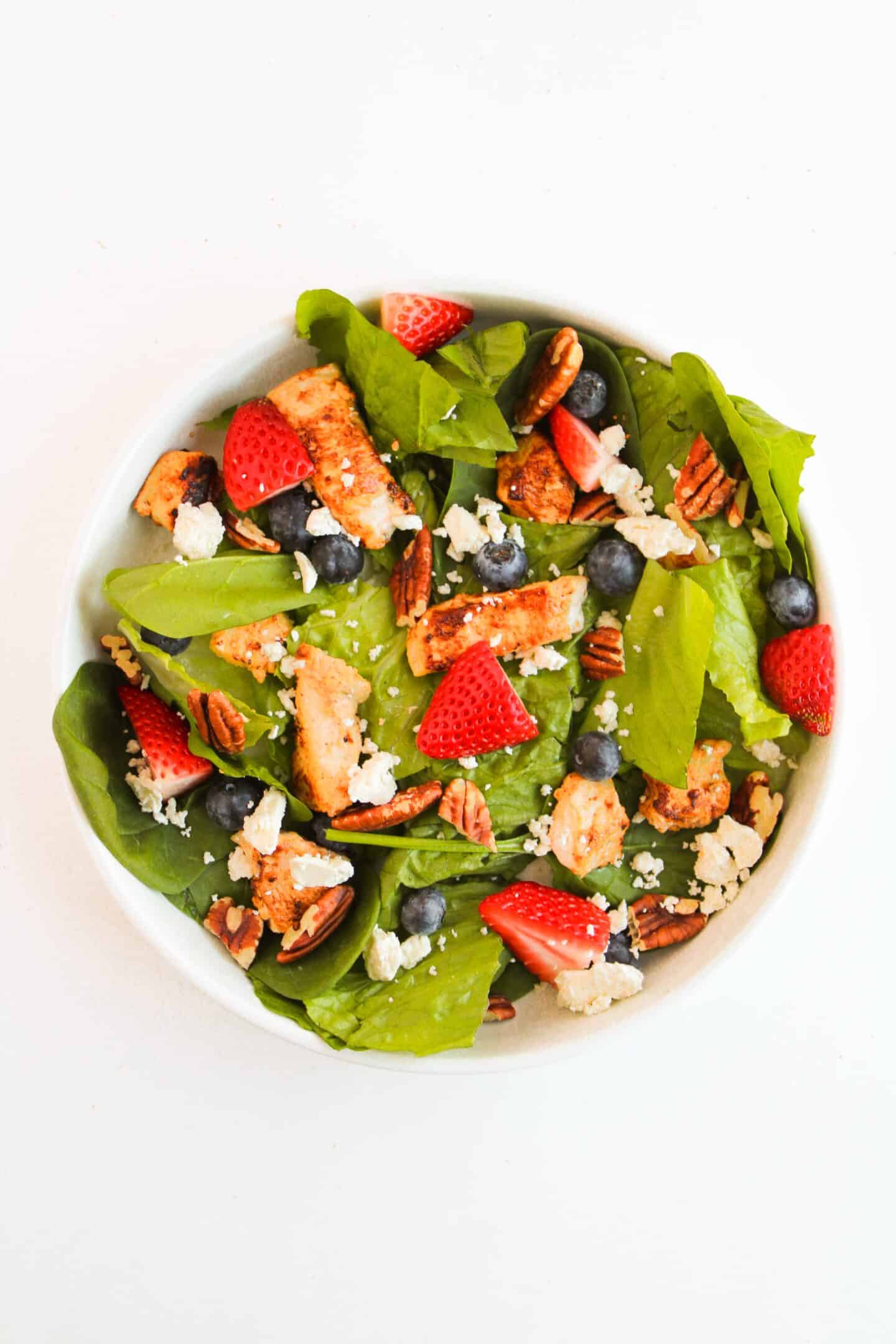 A delicious chicken salad with grilled chicken pieces, spinach leaves, strawberries, blueberries, pecans, and crumbled cheese is perfectly complemented by a creamy Greek yogurt dressing, all served in an elegant bowl on a white background.