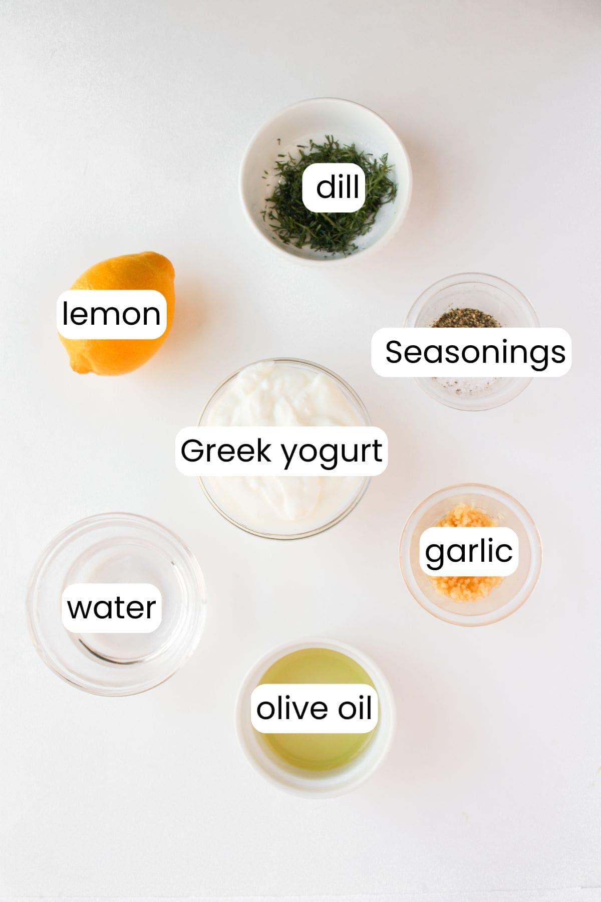 Ingredients for a delicious chicken salad recipe are arranged on a white surface: lemon, dill, seasonings, Greek yogurt for the creamy dressing, olive oil, garlic, and water.