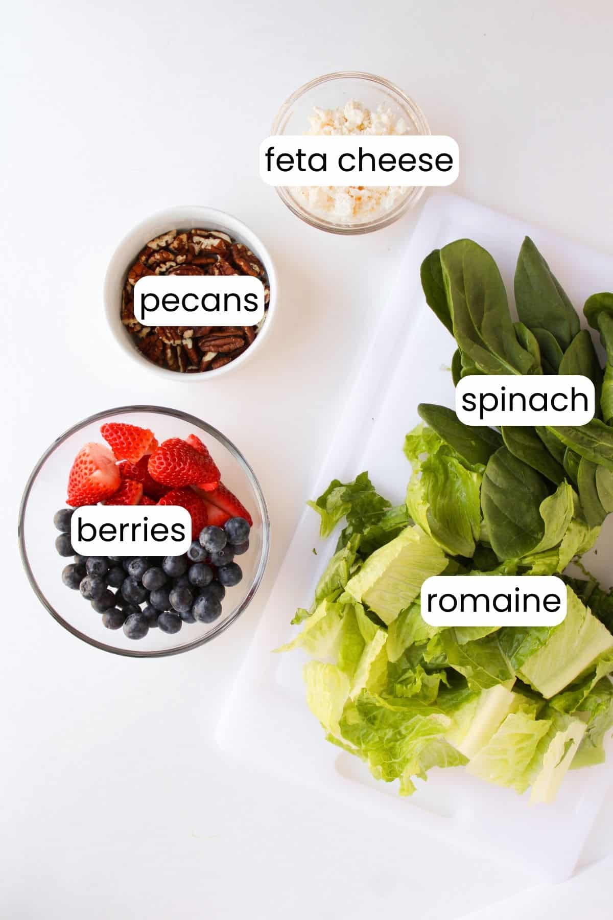 Top-down view of a salad ingredient setup featuring bowls of pecans, feta cheese, berries, and Greek yogurt dressing alongside a cutting board with spinach and romaine lettuce, each labeled accordingly.