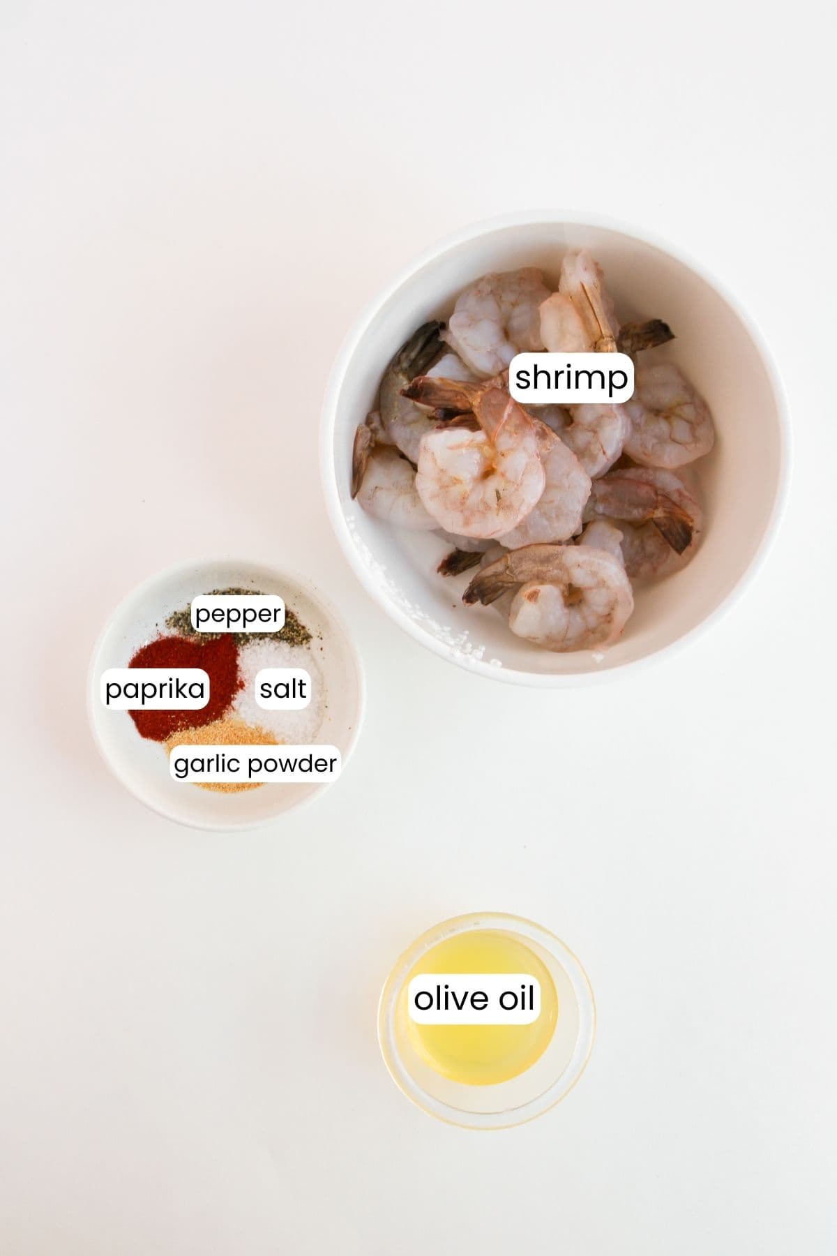 A bowl of succulent shrimp, nestled alongside a vibrant chimichurri mix of spices—pepper, paprika, salt, garlic powder—and a container of olive oil on a light surface.