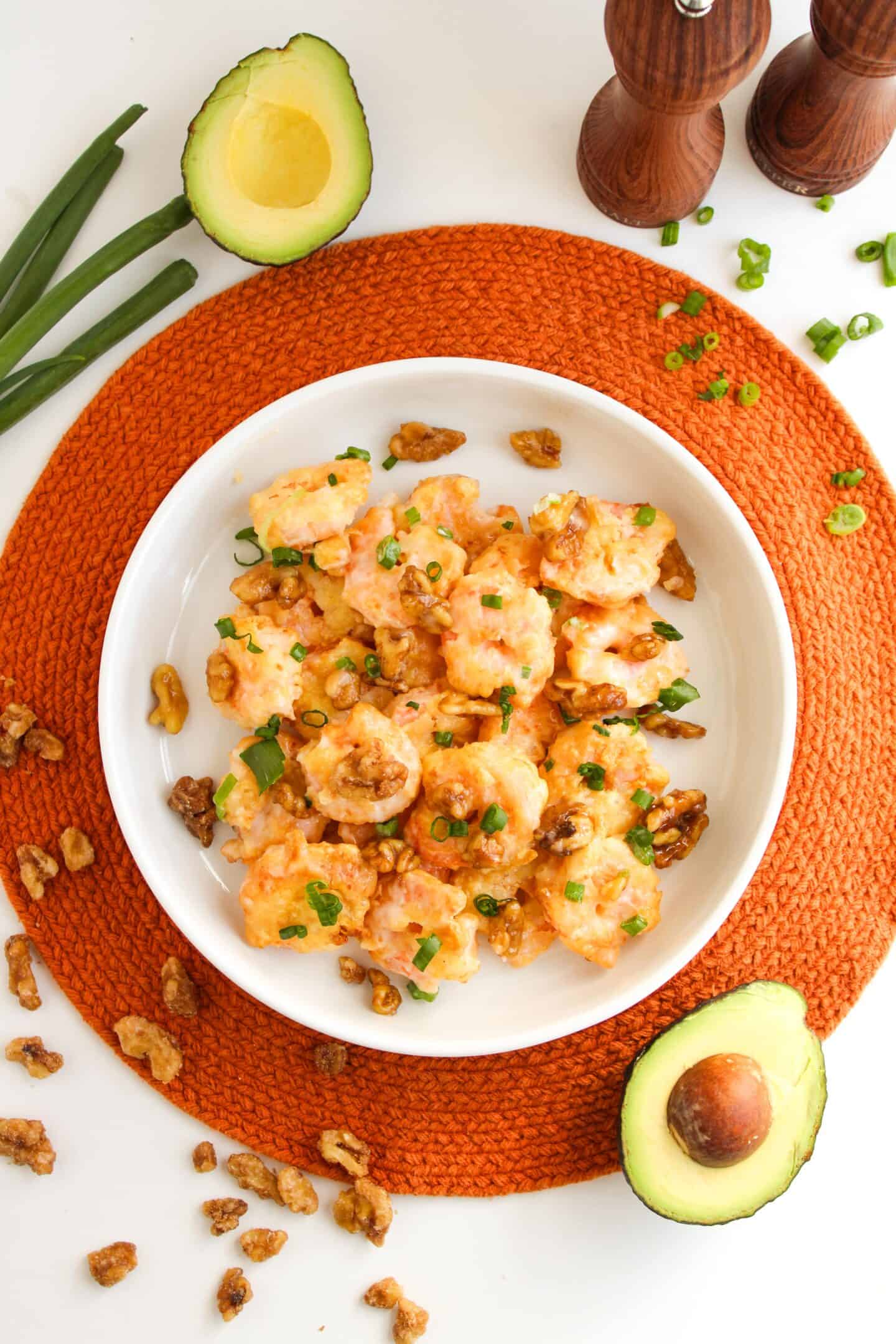 A white bowl of honey walnut shrimp salad, topped with chopped green onions and walnuts, sits on an orange placemat. Nearby, avocado halves and green onions wait to be included—perfect for a fresh twist on air fryer delights.