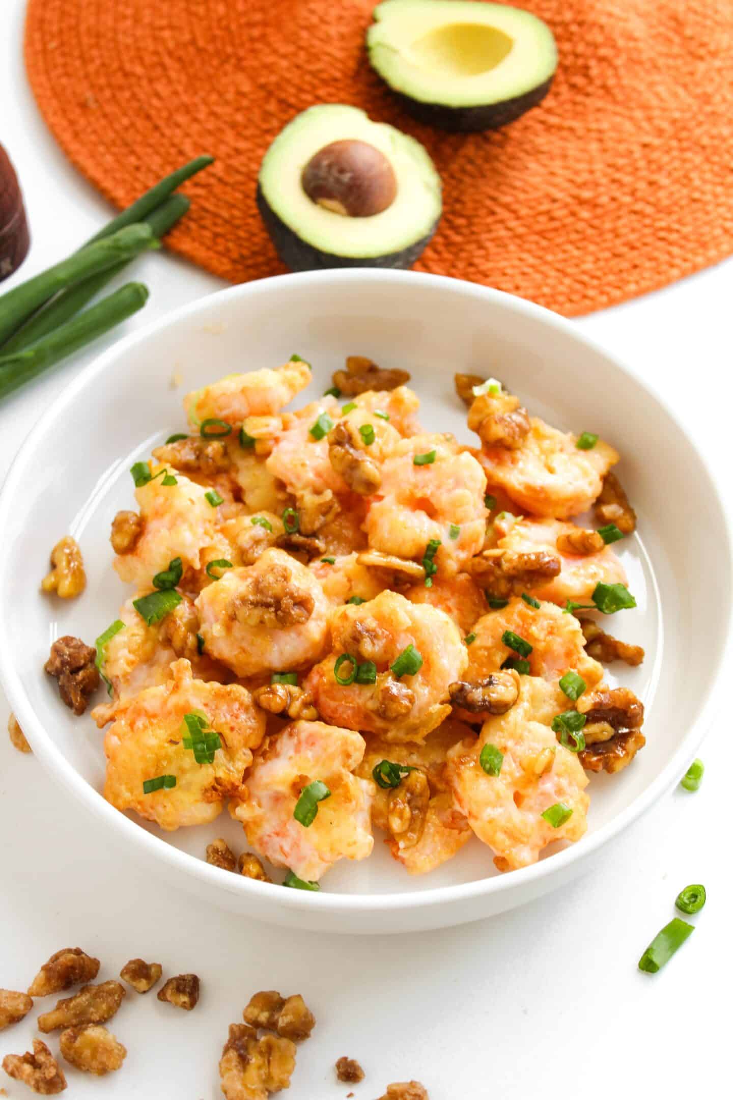 A bowl of Honey Walnut Shrimp with sauce, topped with chopped green onions and crunchy walnuts. In the background, halved avocado and green onions rest on an orange placemat, hinting at a fresh, vibrant meal easily made in an air fryer.