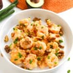 A bowl of Honey Walnut Shrimp with sauce, topped with chopped green onions and crunchy walnuts. In the background, halved avocado and green onions rest on an orange placemat, hinting at a fresh, vibrant meal easily made in an air fryer.