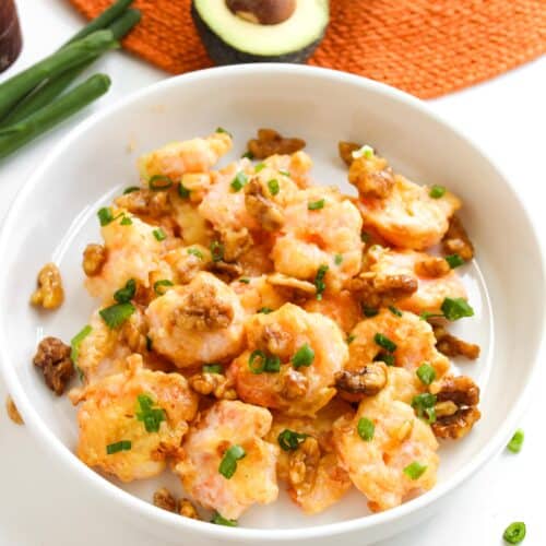 A bowl of Honey Walnut Shrimp with sauce, topped with chopped green onions and crunchy walnuts. In the background, halved avocado and green onions rest on an orange placemat, hinting at a fresh, vibrant meal easily made in an air fryer.