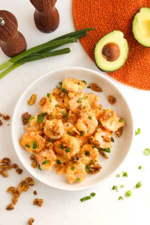 A plate of honey walnut shrimp and walnut salad garnished with chopped green onions, next to whole avocados, green onions, and salt and pepper shakers on a white surface with an orange mat.