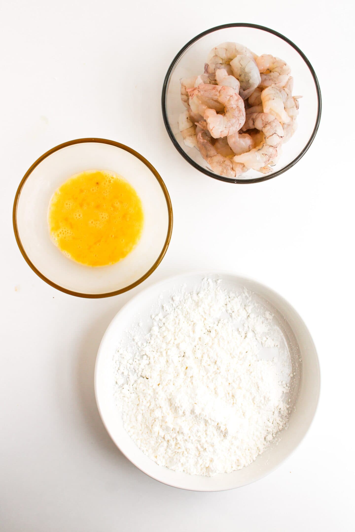 Bowls containing raw shrimp, beaten egg, and flour sit on a white surface, ready to create delicious honey walnut shrimp in your air fryer.