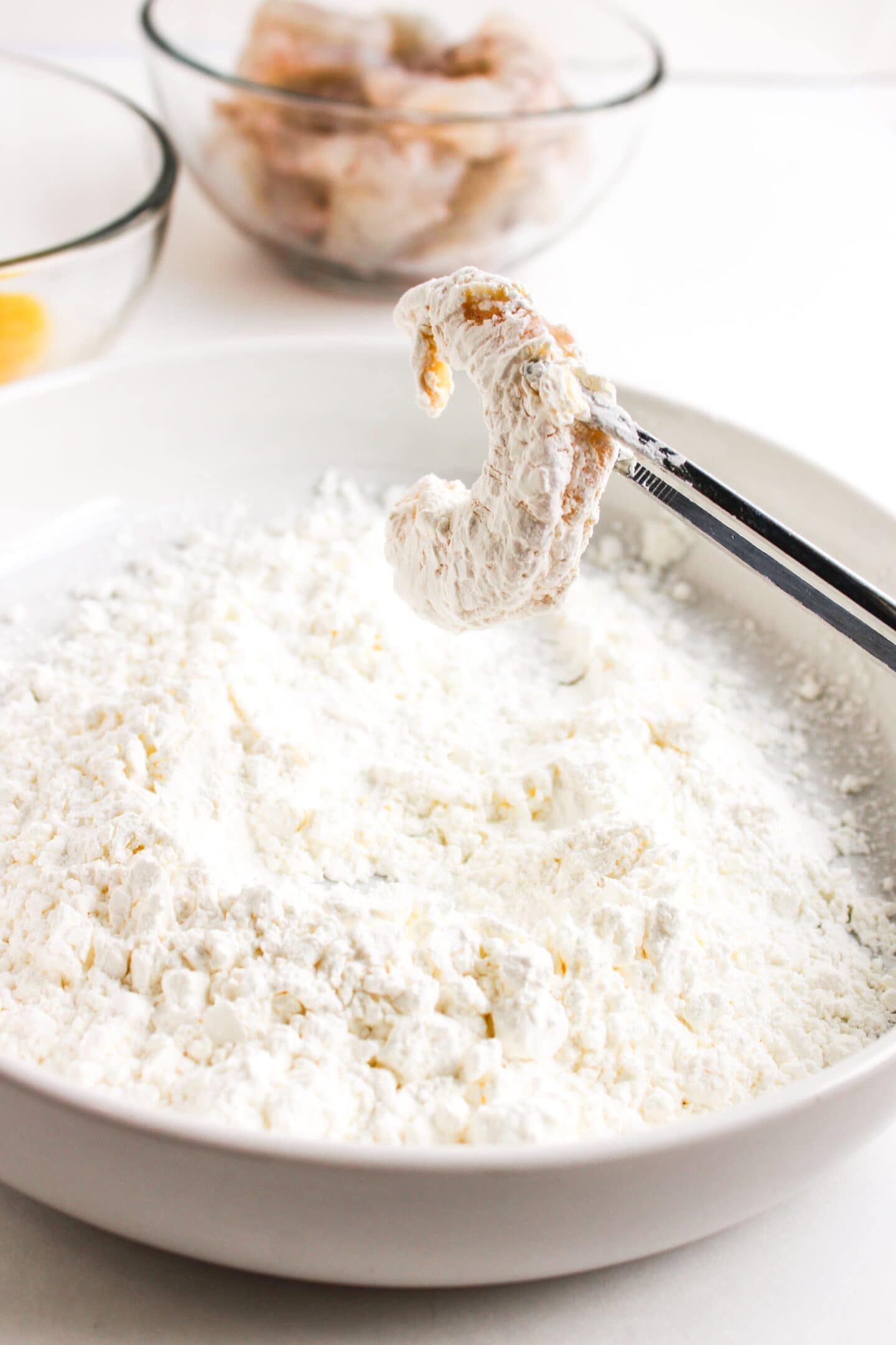 A piece of shrimp is held by tongs over a bowl of white flour, poised for its transformation into a crispy delight. In the background, two bowls await with shrimp and eggs—preparing for an air fryer twist on honey walnut shrimp.