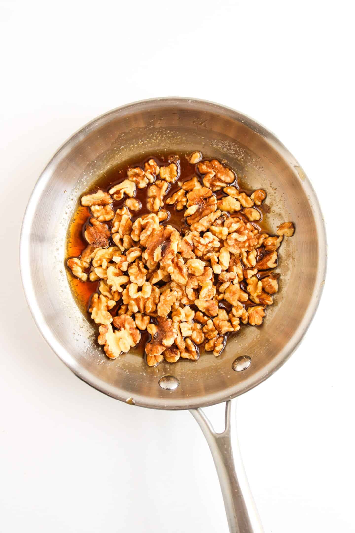 Walnuts being toasted in a stainless steel pan on a white surface, ready to add to your delectable Honey Walnut Shrimp dish.
